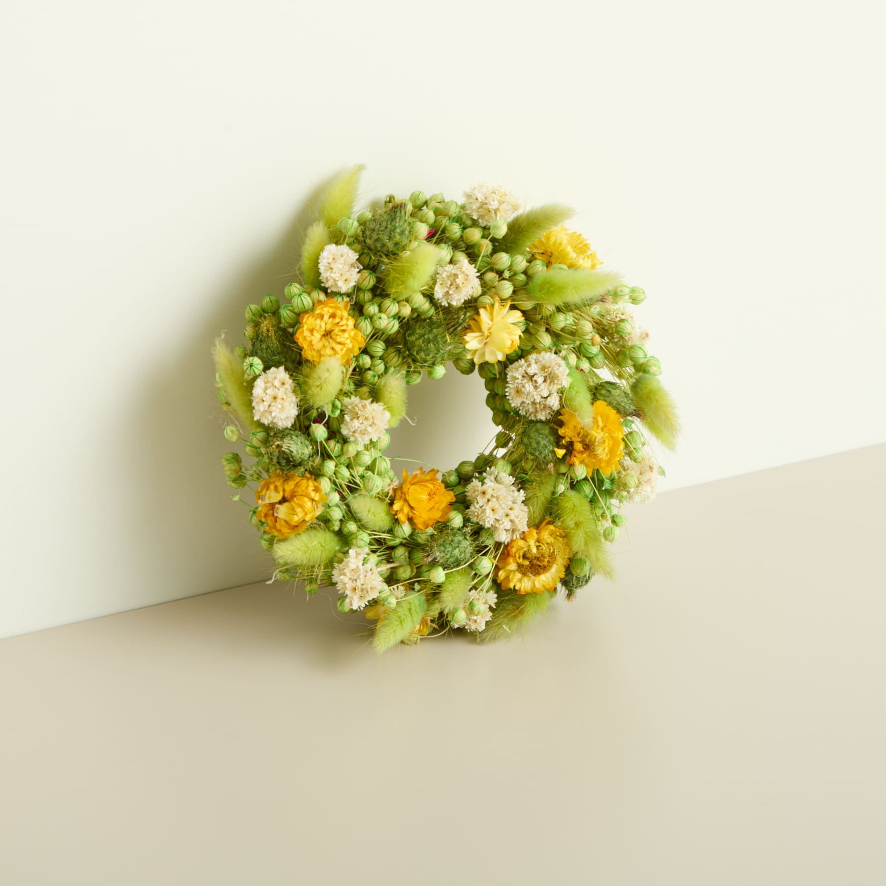 Ein leuchtender Kranz aus grünen und gelben Blumen, Blättern und anderen natürlichen Elementen befindet sich auf einem schlichten, hellen Hintergrund.