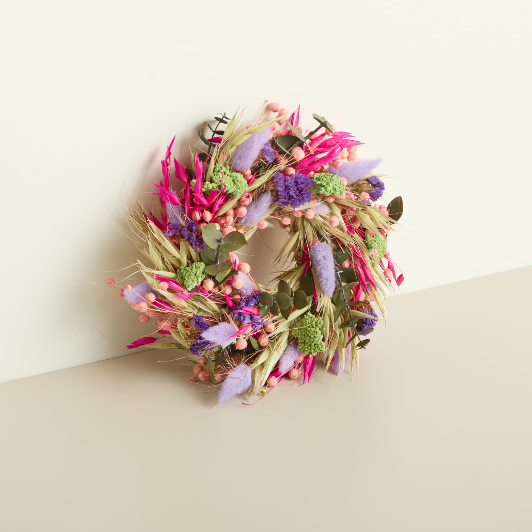 Ein lebendiger, farbenfroher Kranz aus verschiedenen getrockneten Blumen, Blättern und anderen natürlichen Elementen wird vor einem schlichten, hellen Hintergrund präsentiert.