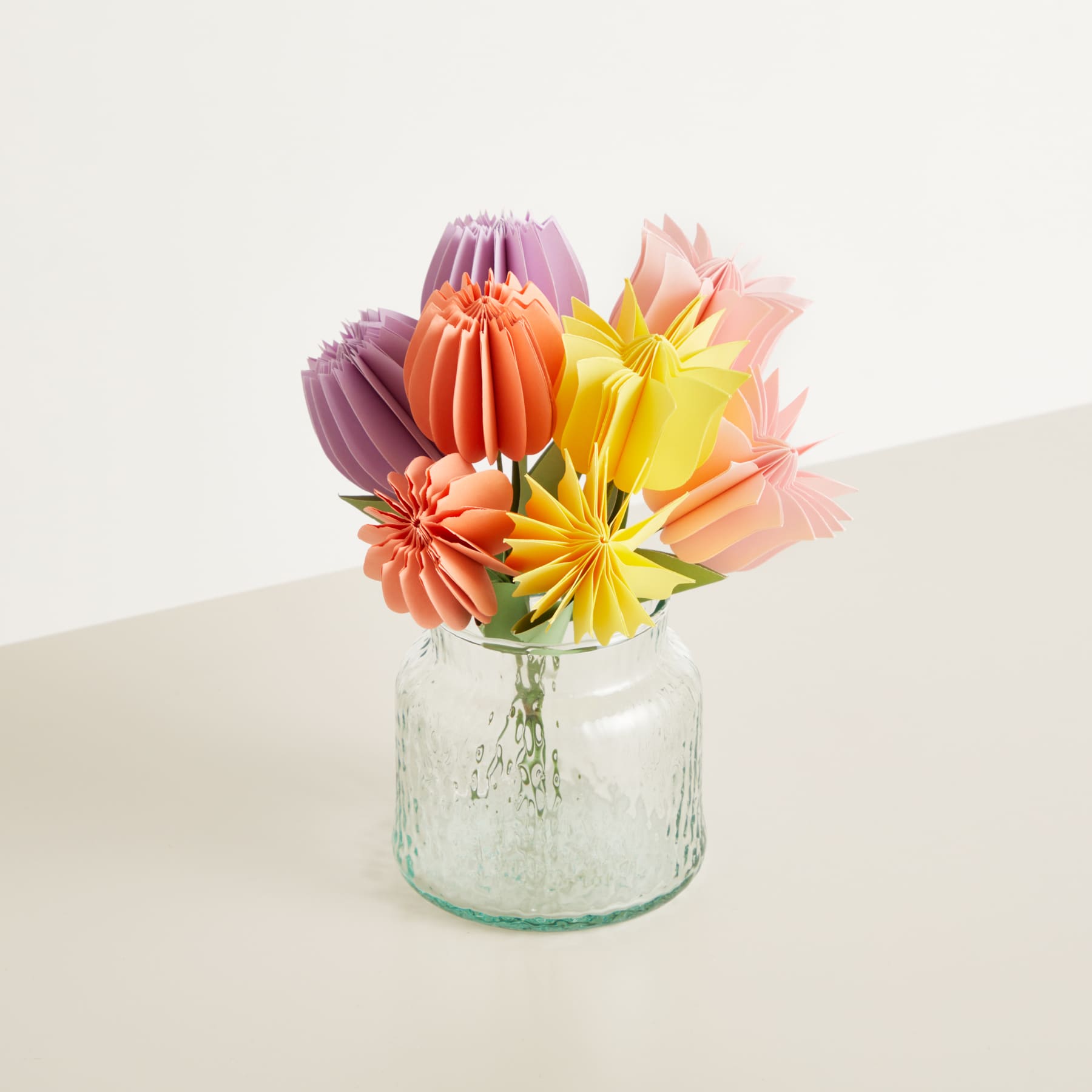 Ein Strauß leuchtender, origamiartiger Papierblumen in Rosa-, Orange- und Gelbtönen wird in einem durchsichtigen Glasgefäß vor einem schlichten, hellen Hintergrund präsentiert.