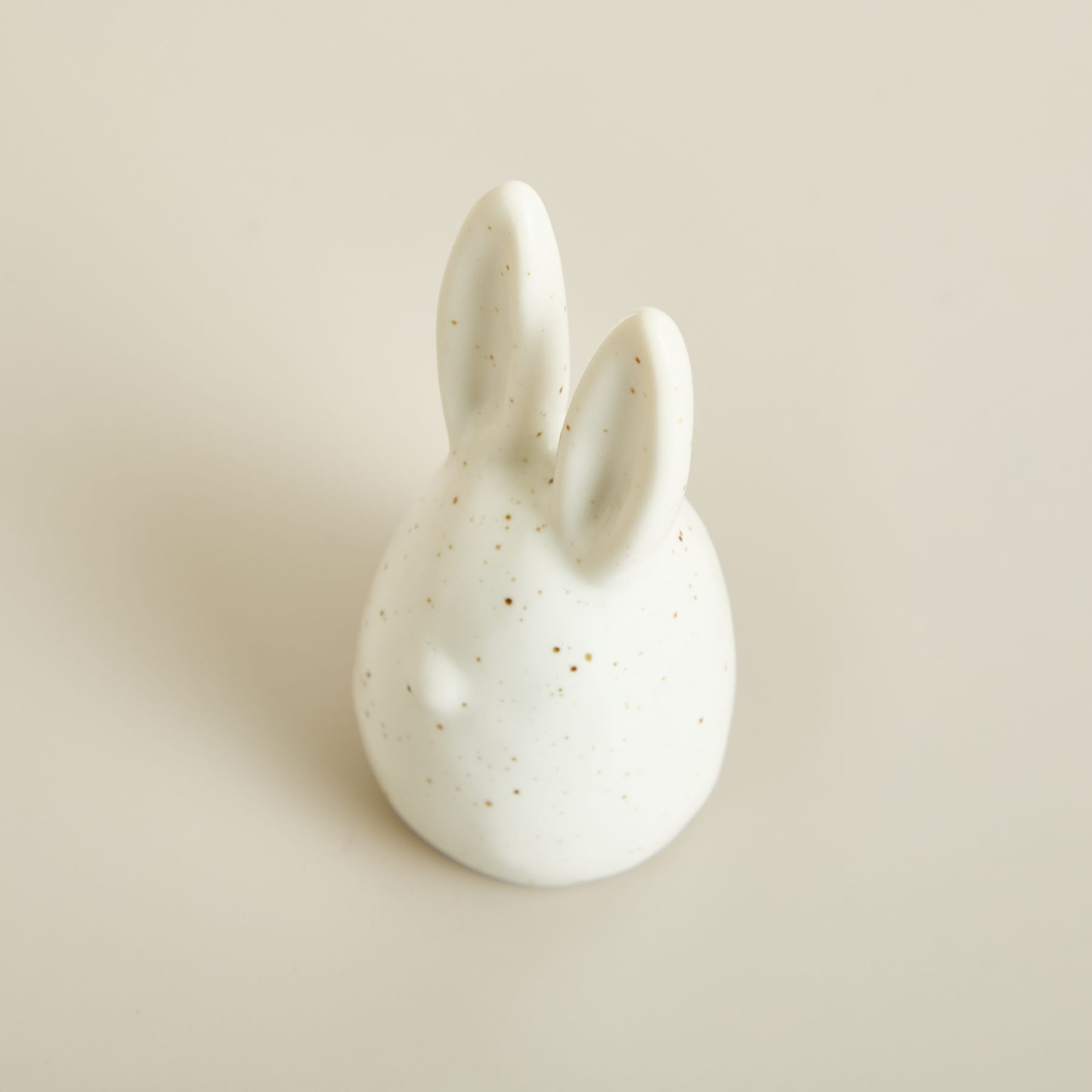 Eine kleine, weiße Keramikfigur, die einem Kaninchen oder Häschen ähnelt, sitzt auf einem schlichten, beigen Hintergrund.