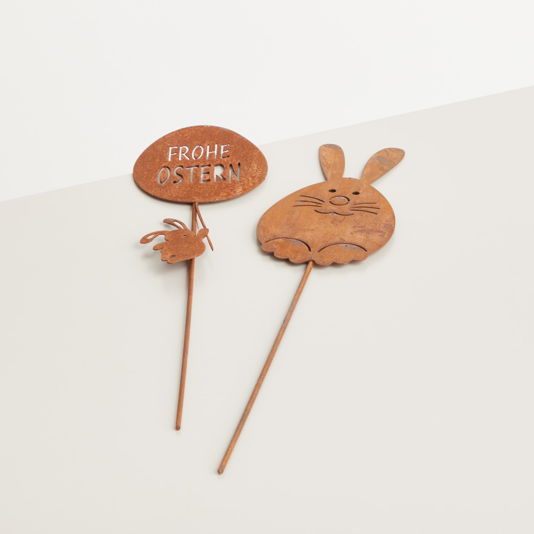 Das Bild zeigt zwei hölzerne Osterdekorationen – eine pilzförmige mit dem deutschen Text „Frohe Ostern“ und eine hasenförmige, beide auf Holzstäben vor einem schlichten weißen Hintergrund montiert.