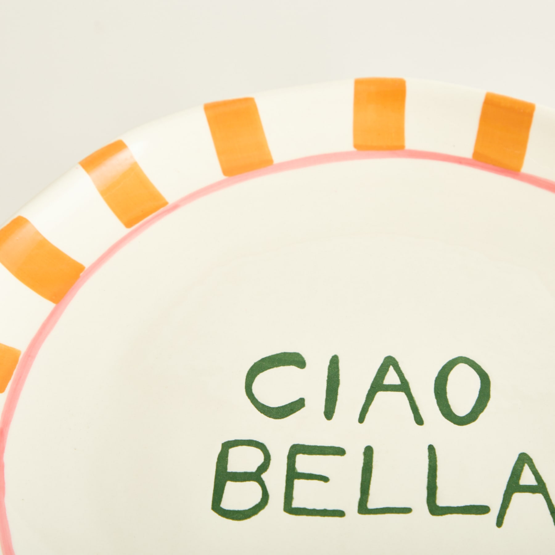Das Bild zeigt einen weißen Teller mit dem Text „CIAO BELLA“ in grünen Buchstaben, umgeben von orangefarbenen und rosa Streifen oder Strahlen.
