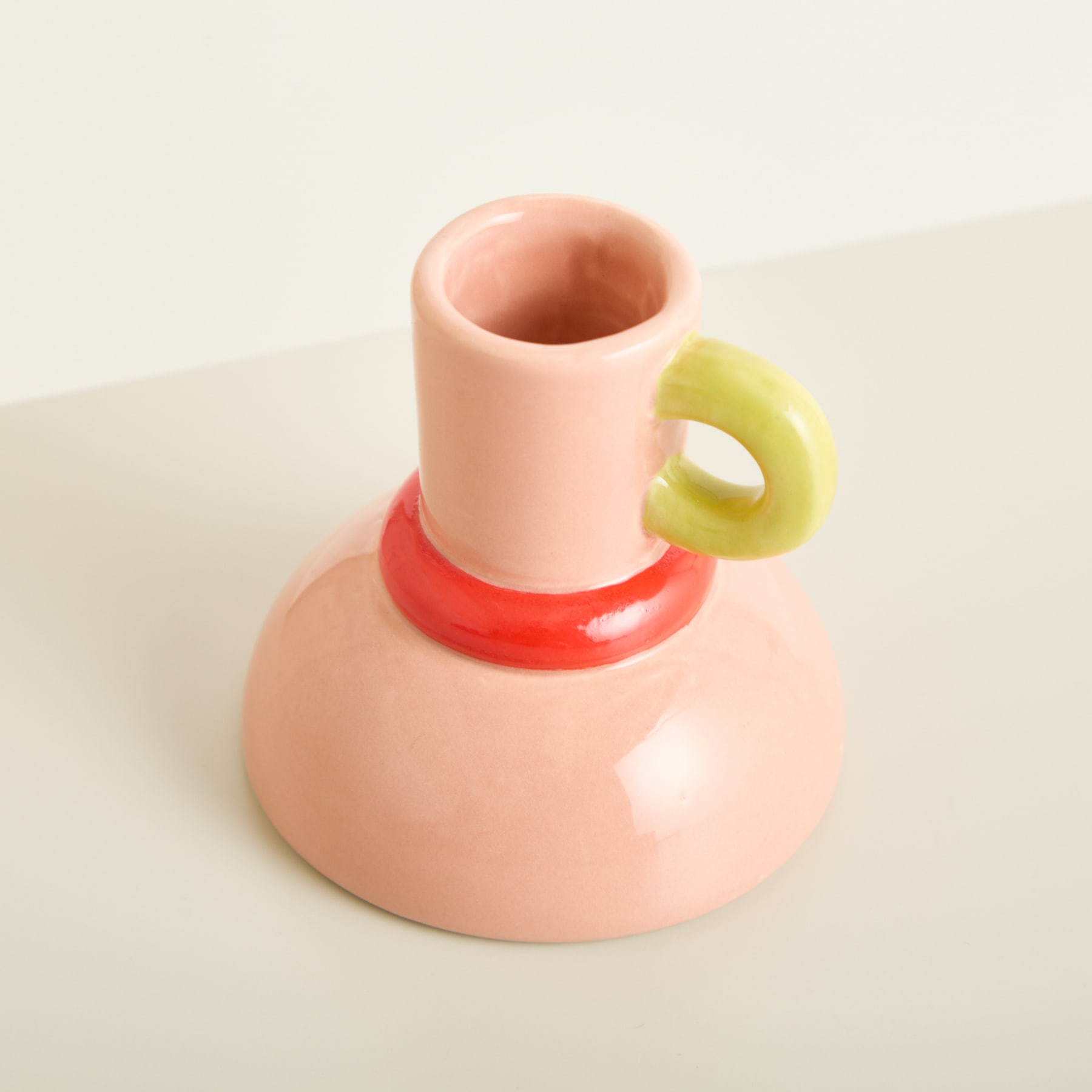 Eine rosafarbene Keramikvase mit einem gelben ringförmigen Griff steht auf einer schlichten weißen Oberfläche.