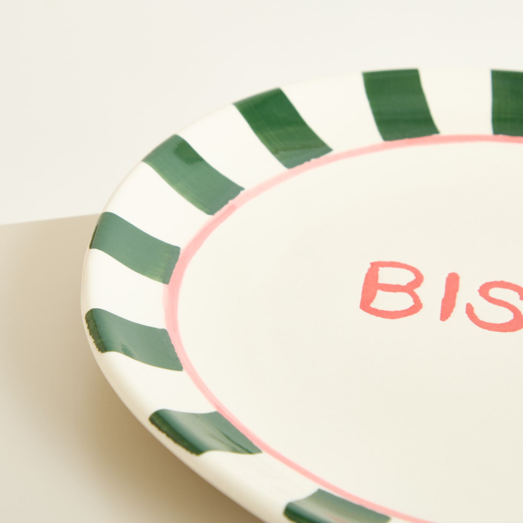 Ein weißer Keramikteller mit grünem und rosa Streifenmuster, in dessen Mitte das Wort „Bis“ in roter Schrift steht.