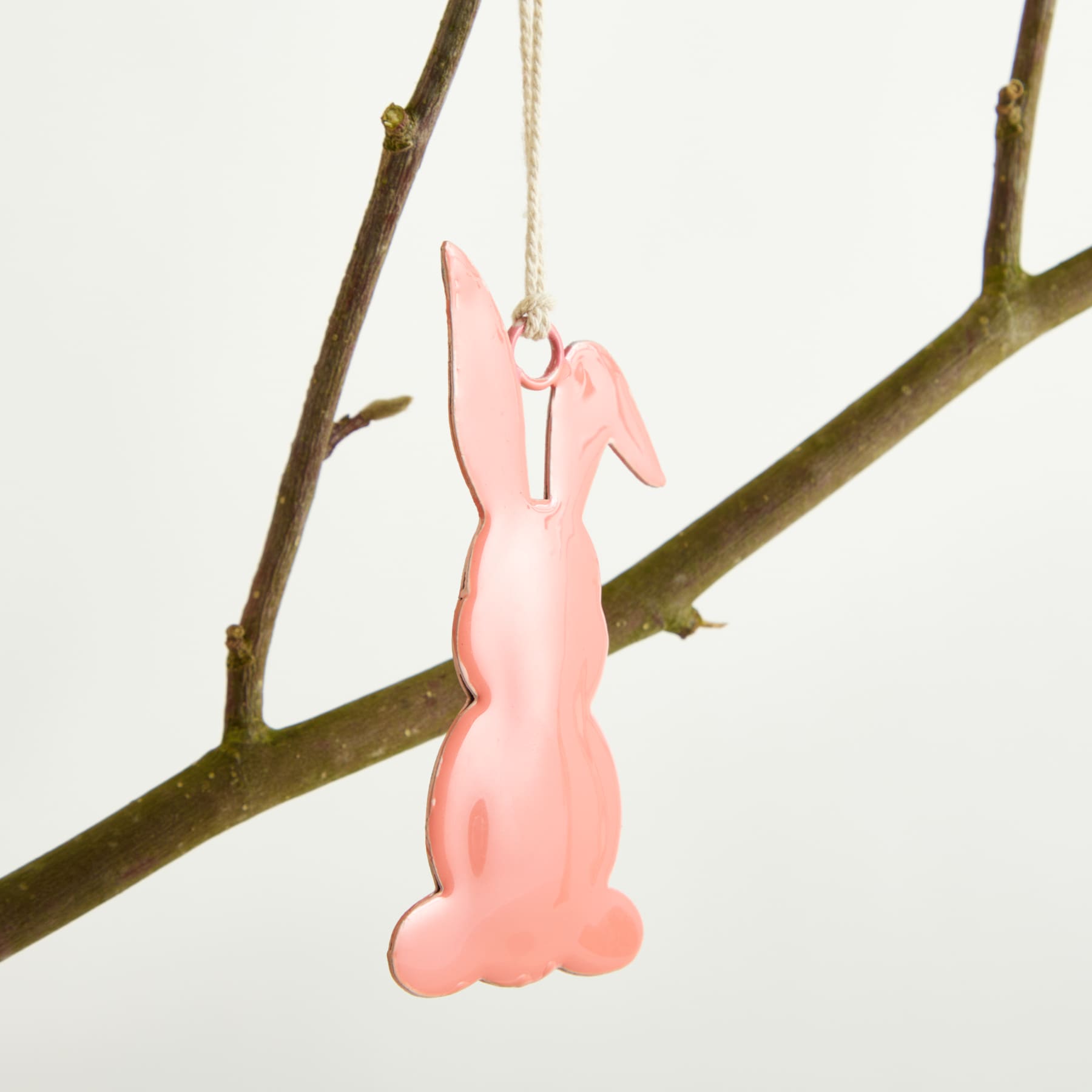 Vor einem schlichten weißen Hintergrund hängt ein rosafarbenes Kaninchen-Ornament aus Holz an einem Ast.