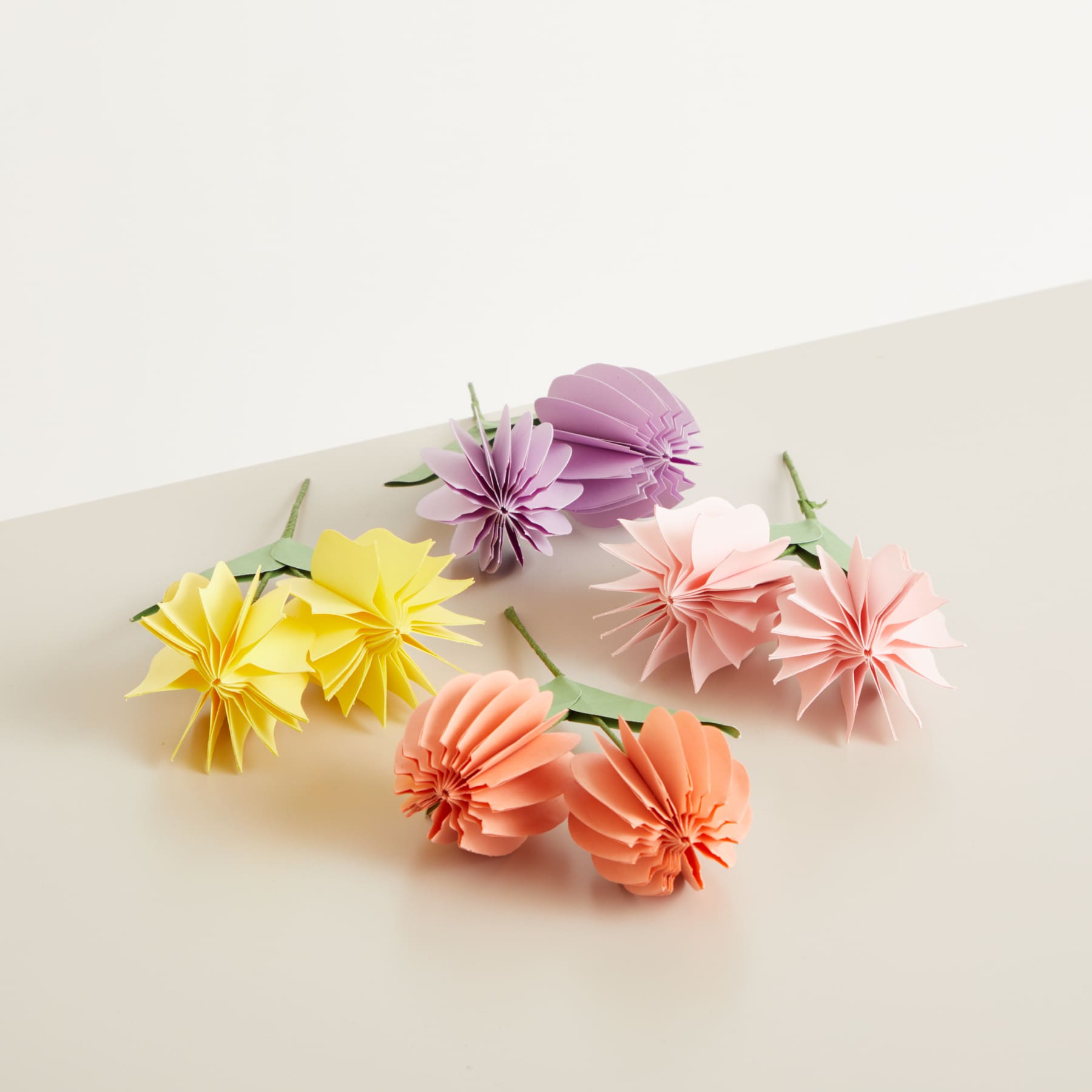 Leuchtende Papierblumen in verschiedenen Pastelltönen, darunter Gelb, Rosa und Lila, sind auf einer schlichten, hellen Oberfläche angeordnet.