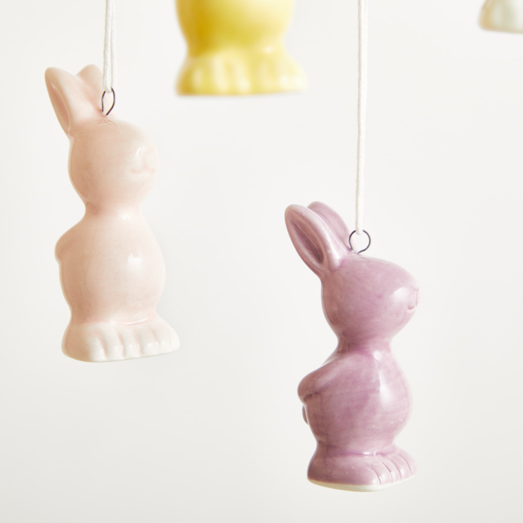 Das Bild zeigt zwei hängende Keramik-Kaninchenfiguren, eine in blassem Rosa und die andere in blassem Gelb, vor einem schlichten weißen Hintergrund.