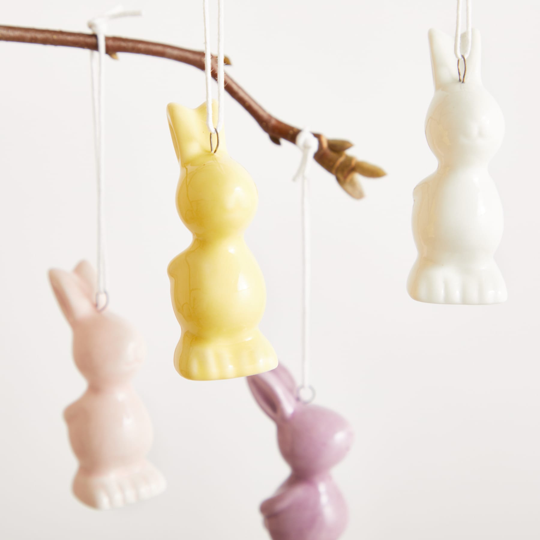 An einem Ast hängen mehrere bunte Kerzen oder Dekorationen in Hasenform in verschiedenen Pastelltönen, darunter Gelb, Rosa und Weiß.