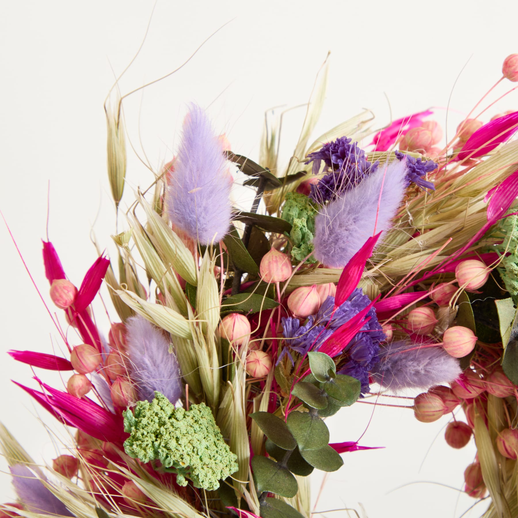 Ein farbenfroher Strauß getrockneter Blumen und Blätter, darunter violette Federn, rosa und grüne Schoten und verschiedene strukturierte Elemente vor einem schlichten weißen Hintergrund.