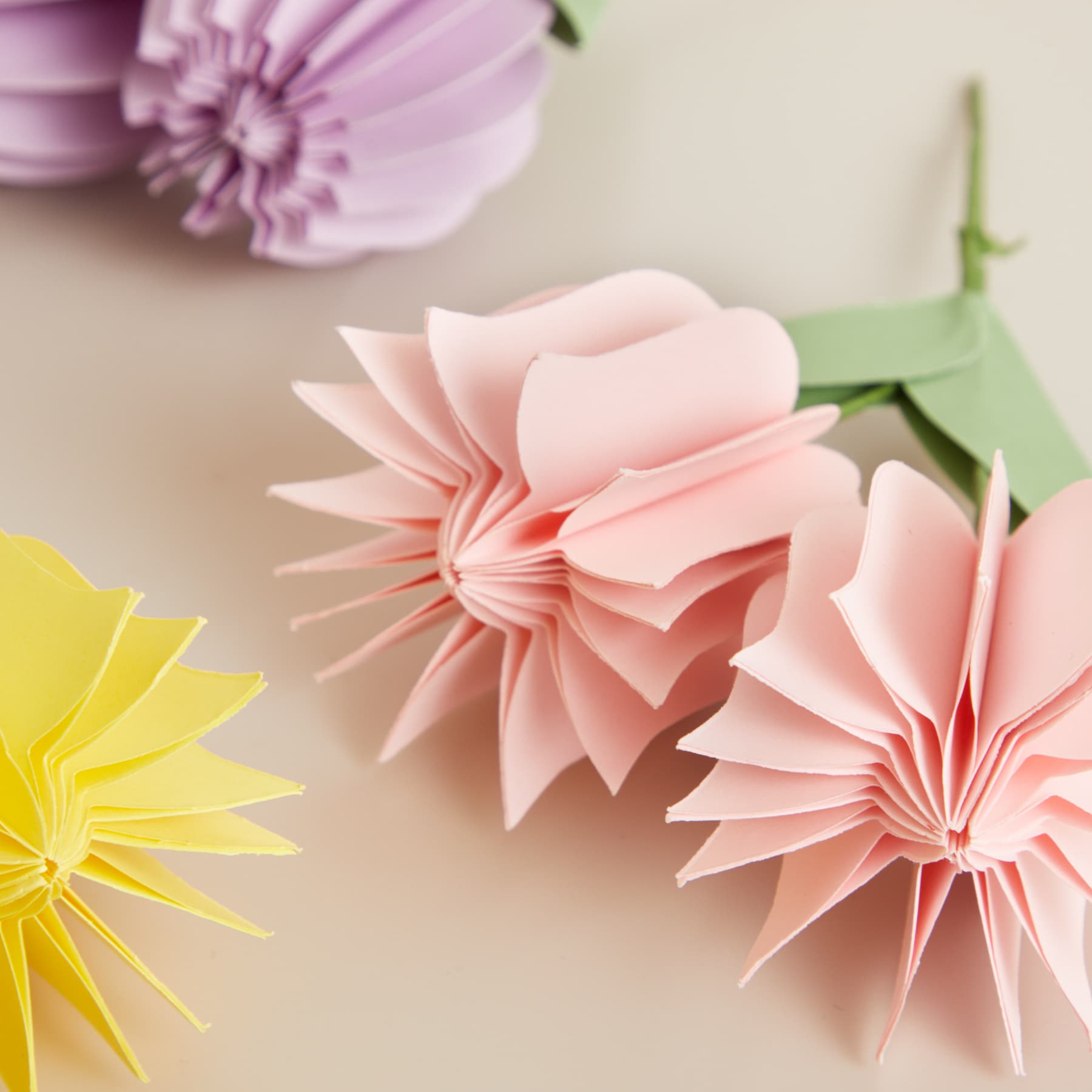 Das Bild zeigt zarte, origamiartige Papierblumen in Rosa- und Gelbtönen vor einem weichen, neutralen Hintergrund.