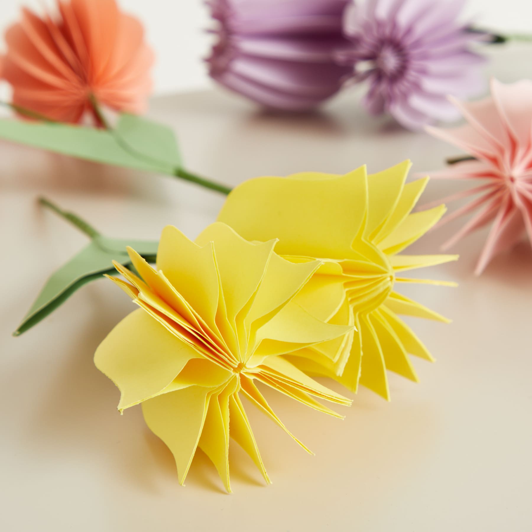 Leuchtende Papierblumen in verschiedenen Farben, darunter Gelb, Orange und Lila, sind auf einer hellen Oberfläche angeordnet.