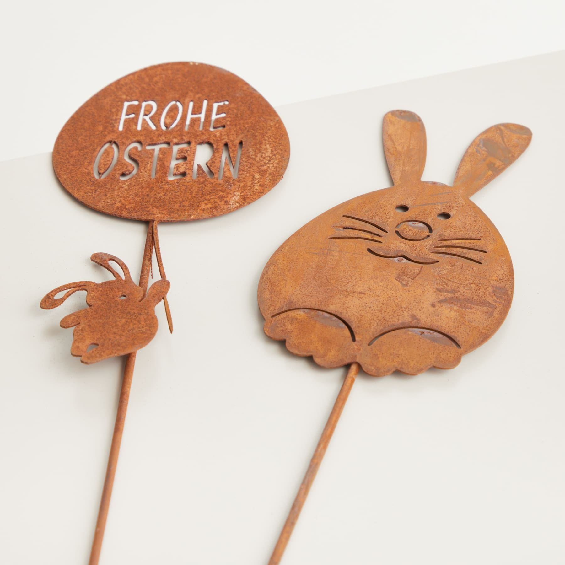 Auf dem Bild sind zwei Deko-Objekte zum Thema Ostern zu sehen – ein Schild mit der Aufschrift „FROHE OSTERN“ und eine Hasenfigur aus Holz.