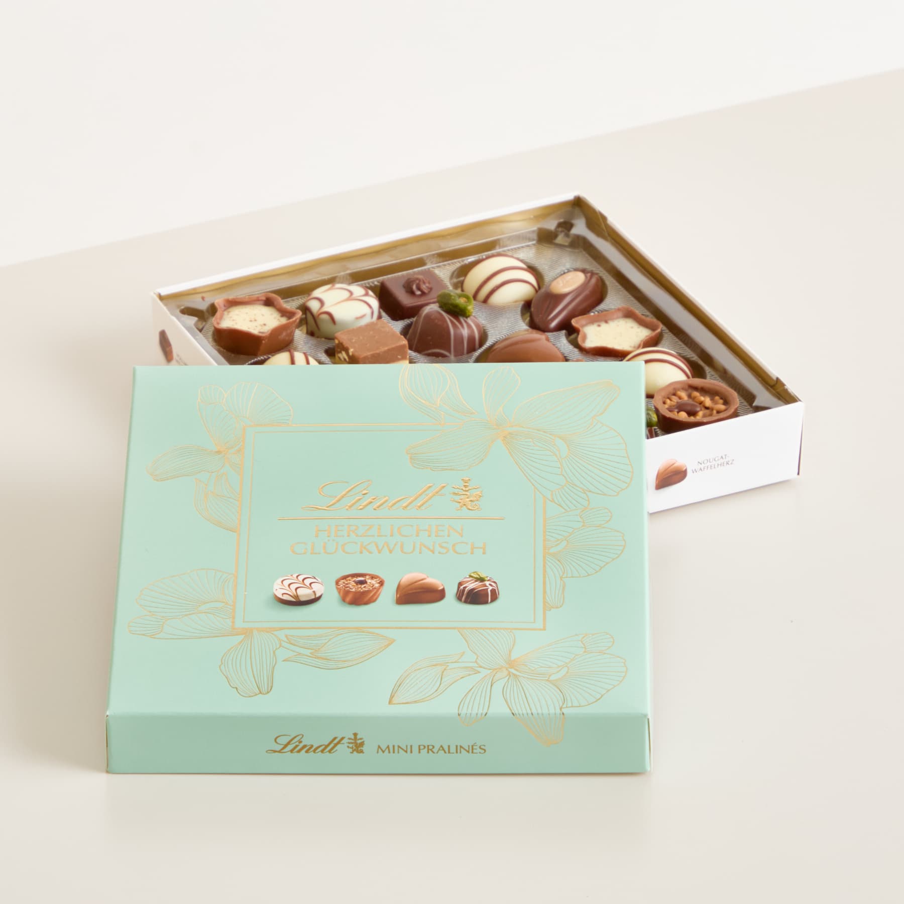 Eine Schachtel mit verschiedenen Lindt-Schokoladen in einer mintgrünen und goldenen Verpackung, die auf einer hellen Oberfläche liegt.