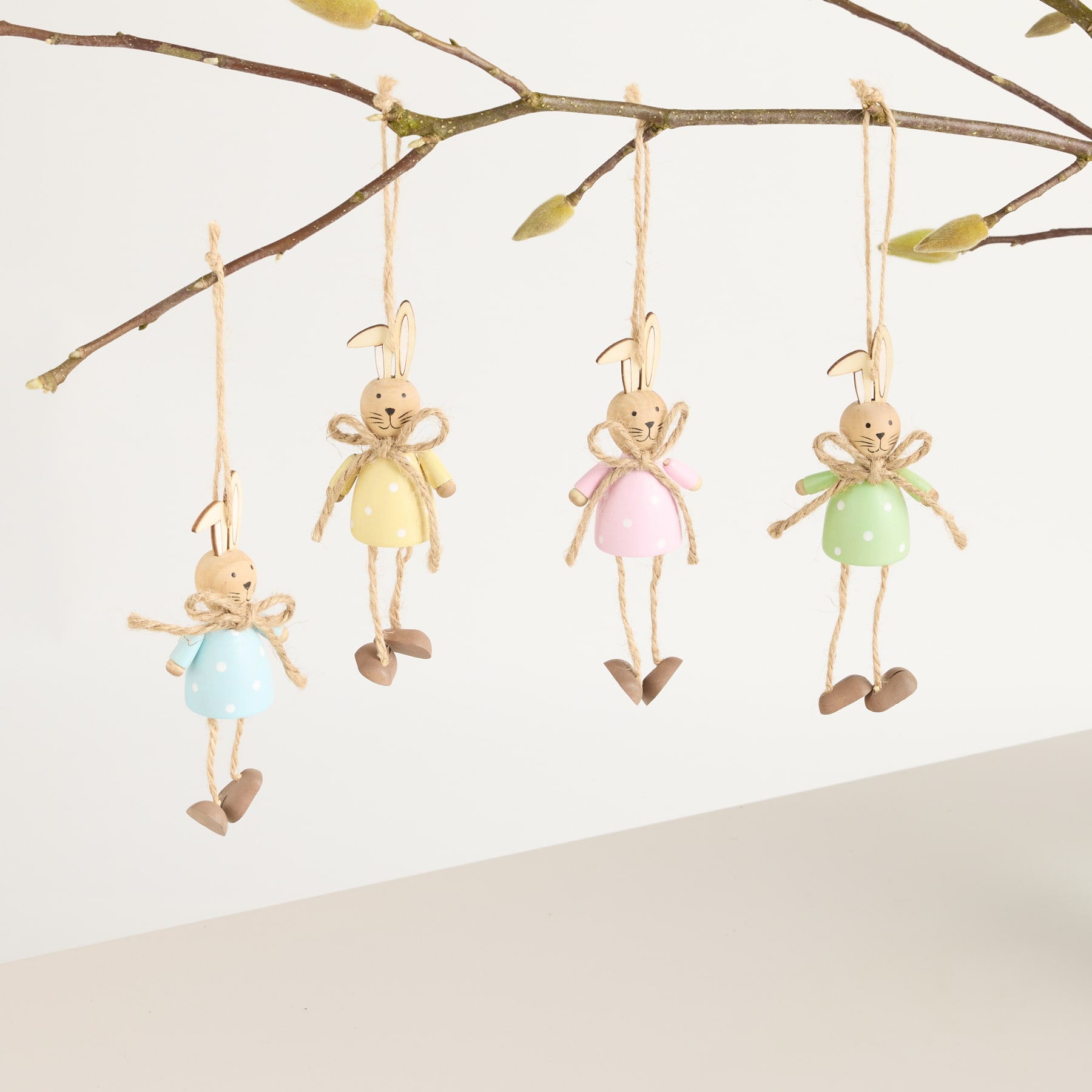 Das Bild zeigt mehrere kleine, skurrile Plüschfiguren, die vor einem schlichten Hintergrund an einem Ast hängen. Die Figuren scheinen stilisierte kaninchen- oder hasenartige Kreaturen in verschiedenen Pastellfarben zu sein, darunter Gelb, Rosa und Grün.