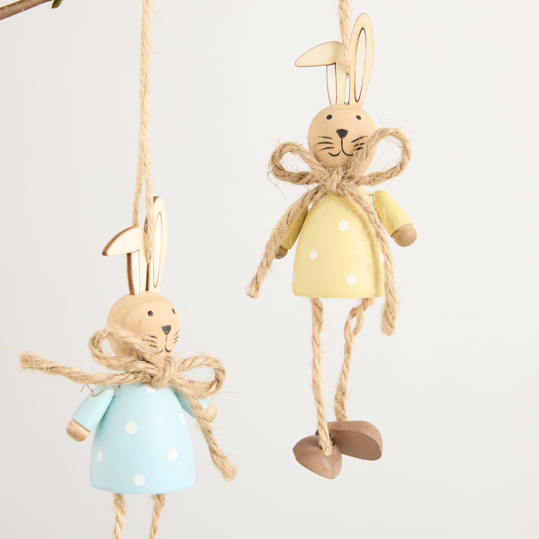 Zwei skurrile Osterhasenfiguren, eine in einem gelben Kleid und die andere in einem blauen Kleid, hängen an Schnüren vor einem schlichten weißen Hintergrund.
