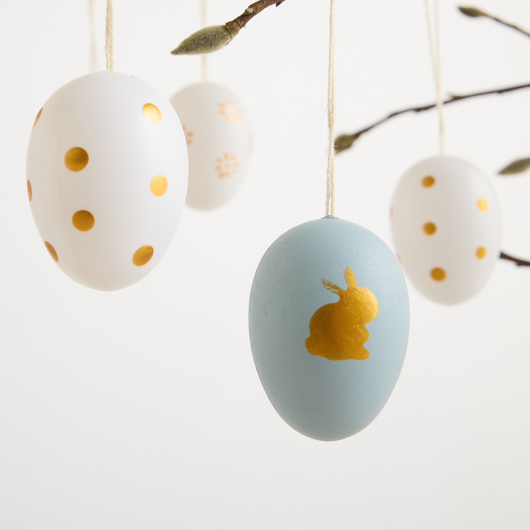 Hängende Ostereier in verschiedenen Pastellfarben, einige mit Tupfen und eines mit der Silhouette eines Hasen, vor einem schlichten weißen Hintergrund.