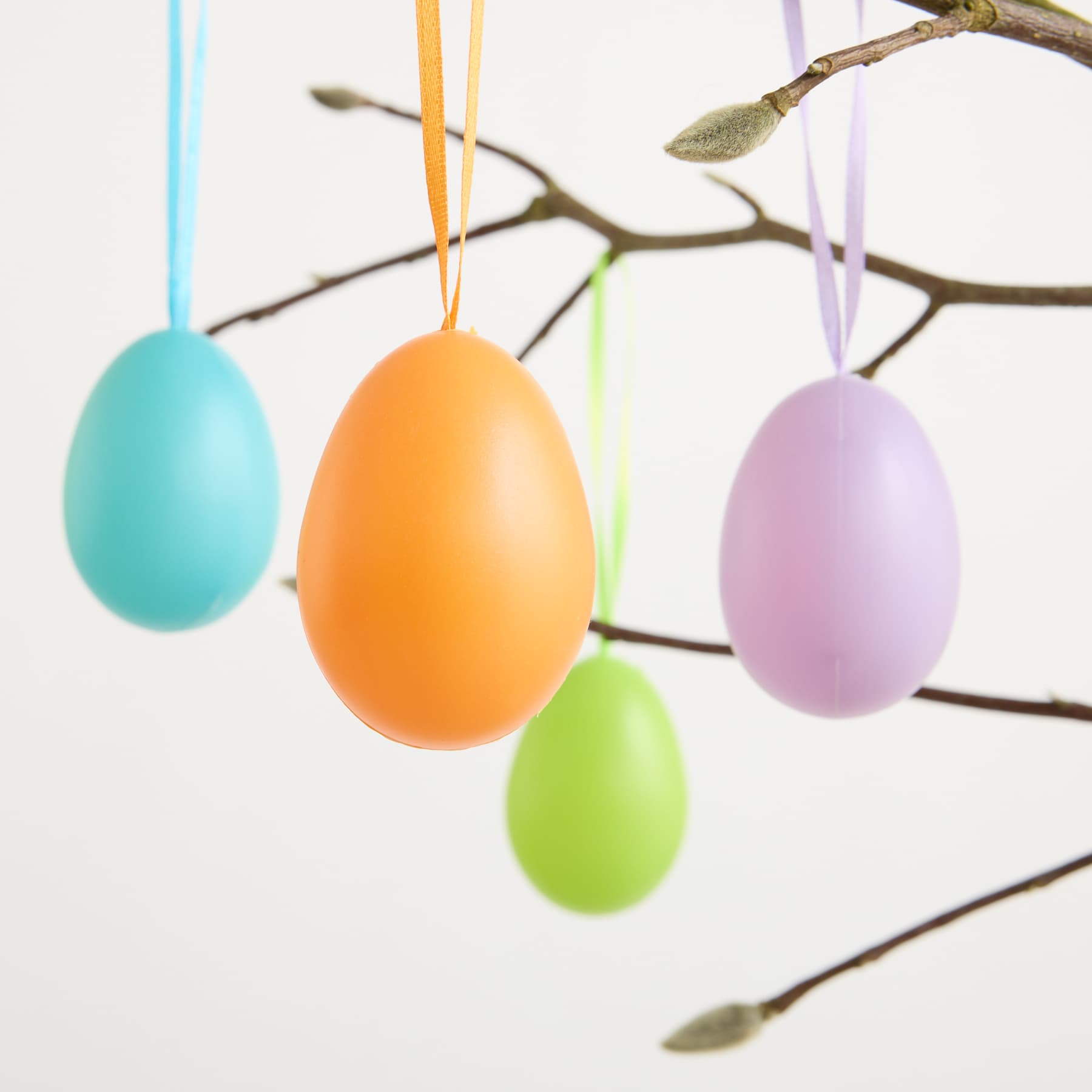 Bunte Ostereier in Pastelltönen aus Blau, Orange, Lila und Grün hängen vor einem schlichten weißen Hintergrund von einem kahlen Ast.