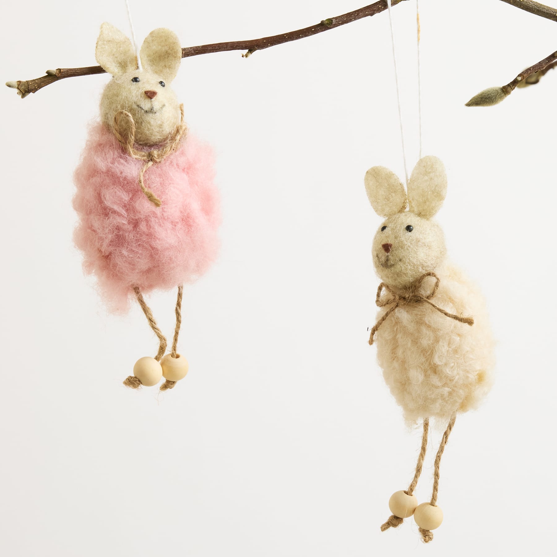 An einem Ast hängen zwei flauschige weiße Kaninchenfiguren, eine trägt einen rosa Stoffbüschel und die andere eine einfache Fliege.
