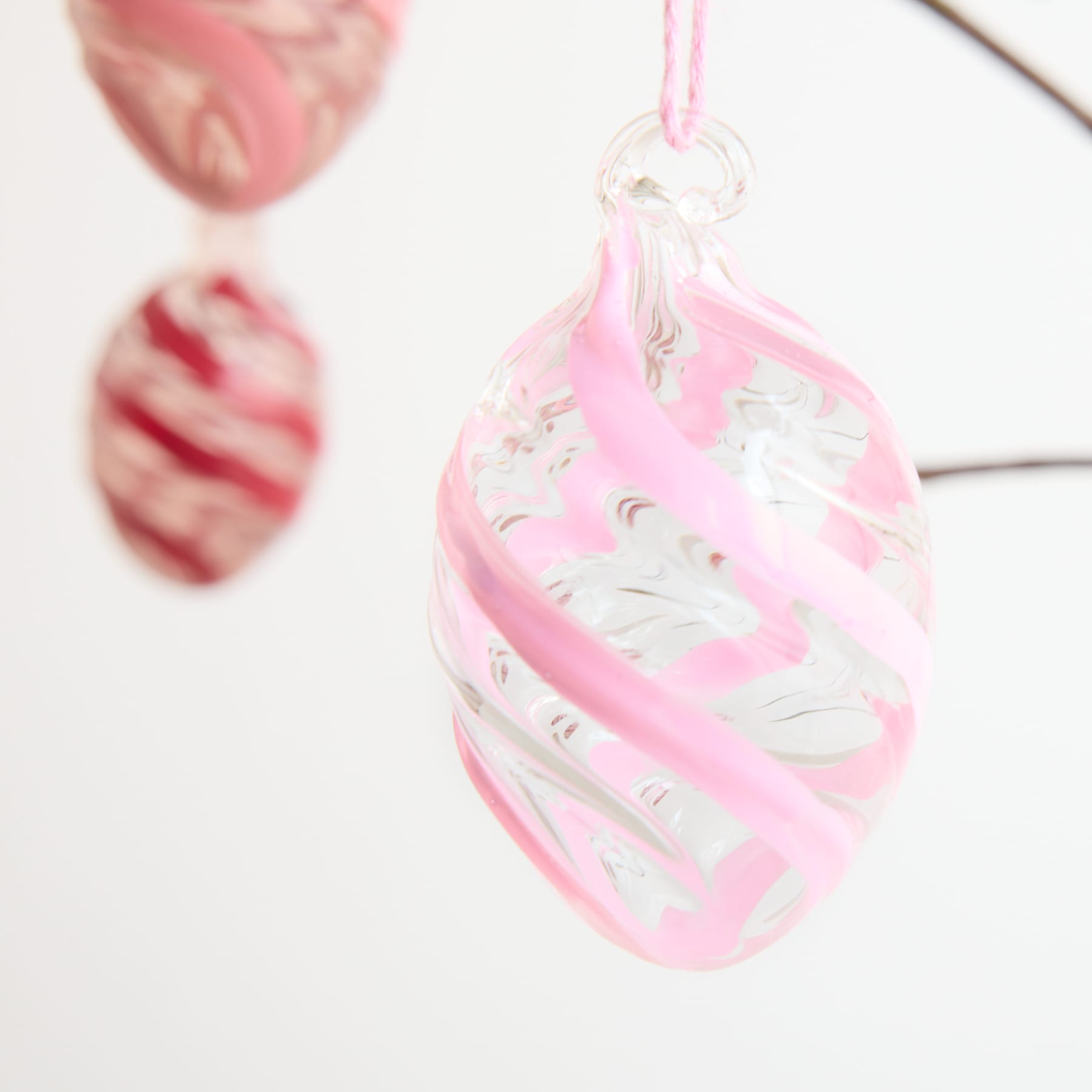 Das Bild zeigt eine Nahaufnahme eines rosa-weiß gewirbelten Glasornaments oder -anhängers mit einer daran befestigten Schnur zum Aufhängen vor einem unscharfen Hintergrund.