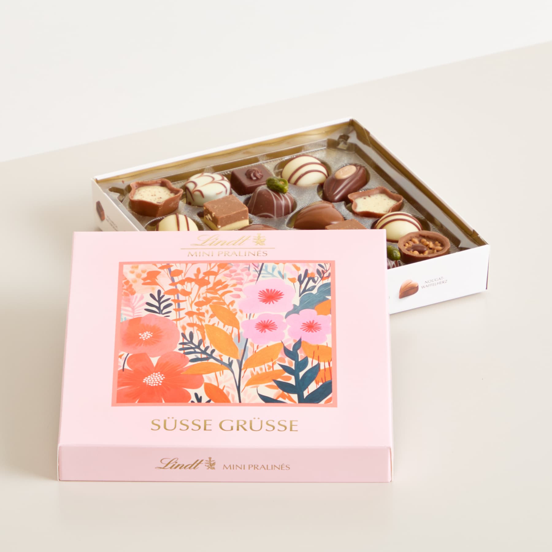 Eine rosa Geschenkbox mit einem Etikett mit Blumenmuster steht auf einem Holztablett, das mit einer Auswahl an Pralinen gefüllt ist.