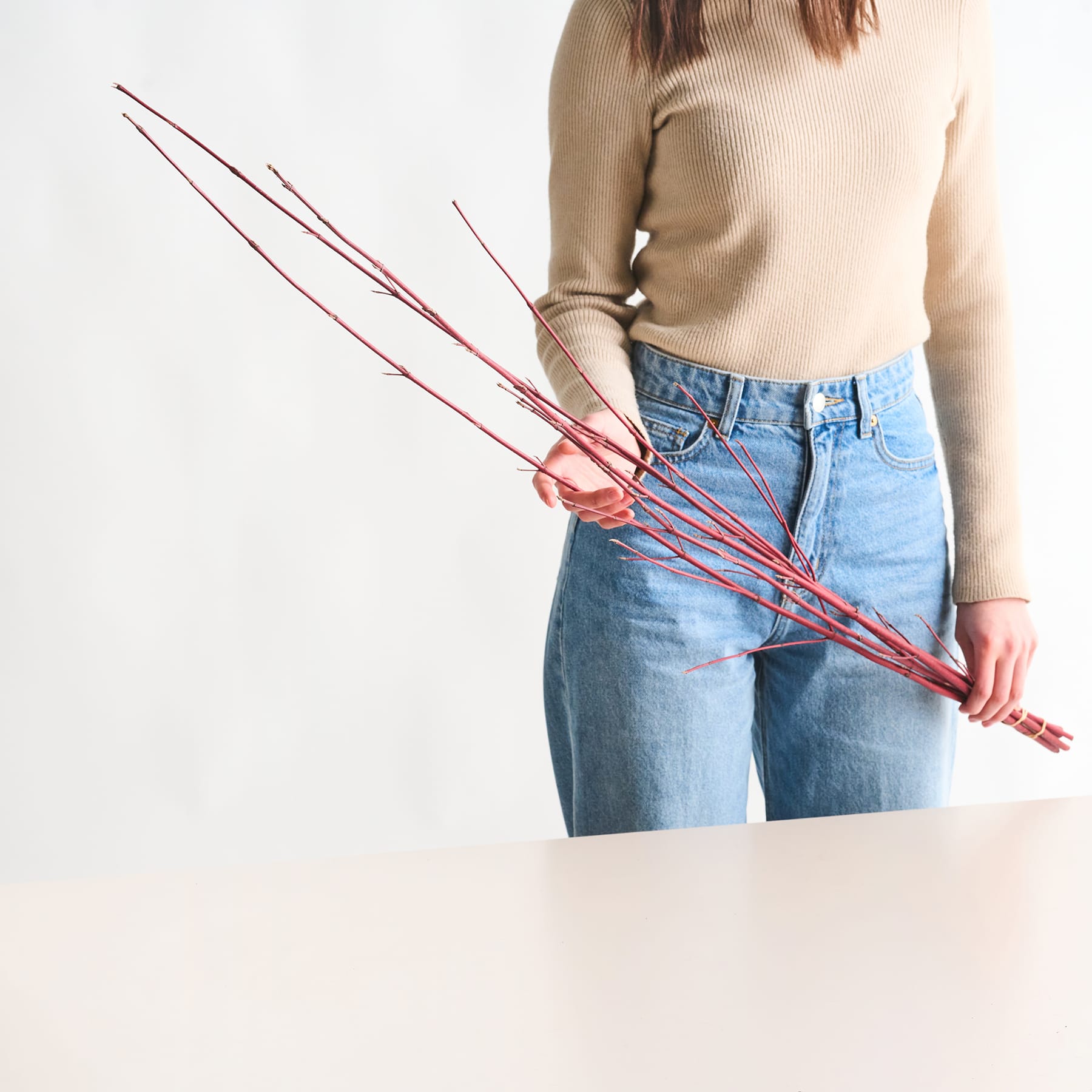Das Bild zeigt eine Person, die einen beigen Pullover und hoch taillierte Bluejeans trägt und vor einem schlichten weißen Hintergrund mehrere rote Essstäbchen in den Händen hält.
