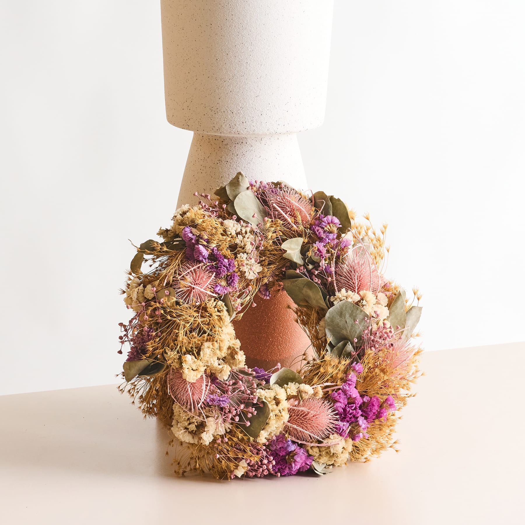 Ein dekorativer Kranz aus getrockneten Blumen und Blättern liegt auf einer Holzoberfläche mit einem schlichten weißen Hintergrund.