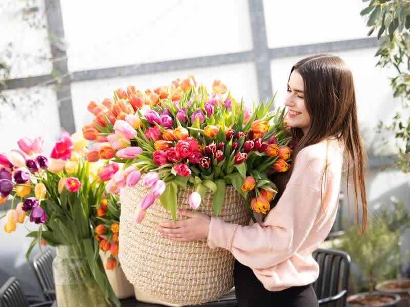 Eine junge Frau mit langem braunem Haar hält einen großen Korb mit einer leuchtend bunten Auswahl an Tulpen und anderen Frühlingsblumen in der Hand und steht in einem hellen und luftigen Raum mit großen Fenstern und Blick auf eine üppige Außenlandschaft.