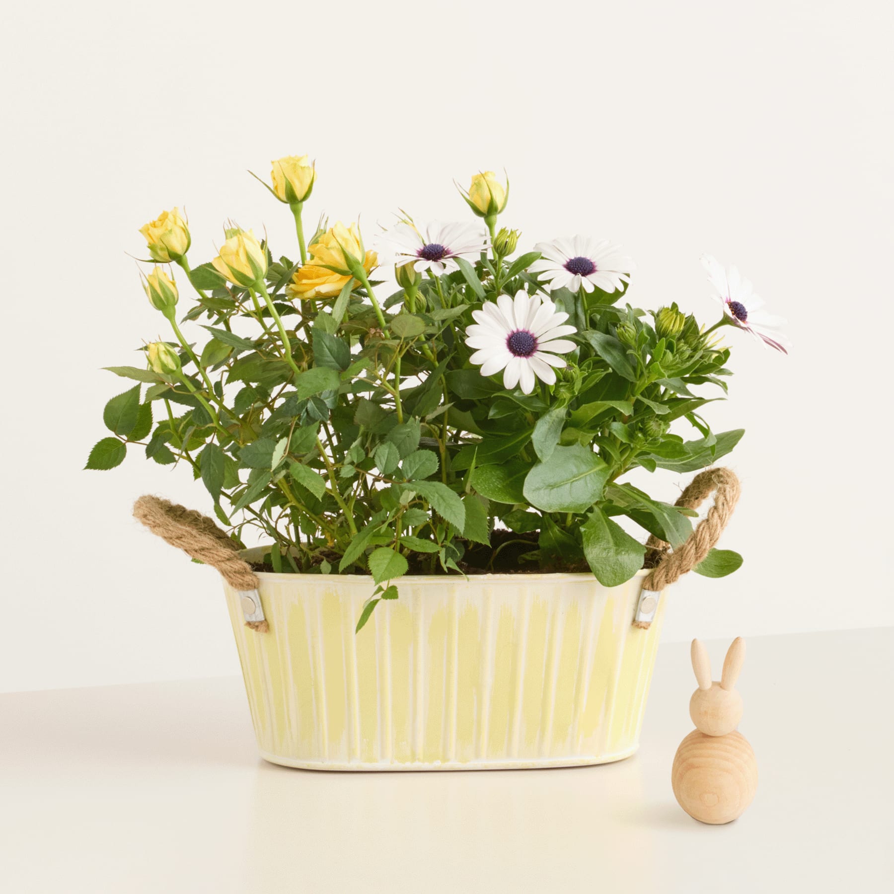 Ein farbenfrohes Arrangement aus gelben Rosen und weißen Gänseblümchen in einem gelben Keramiktopf, begleitet von einer kleinen hölzernen Hasenfigur auf schlichtem Hintergrund.