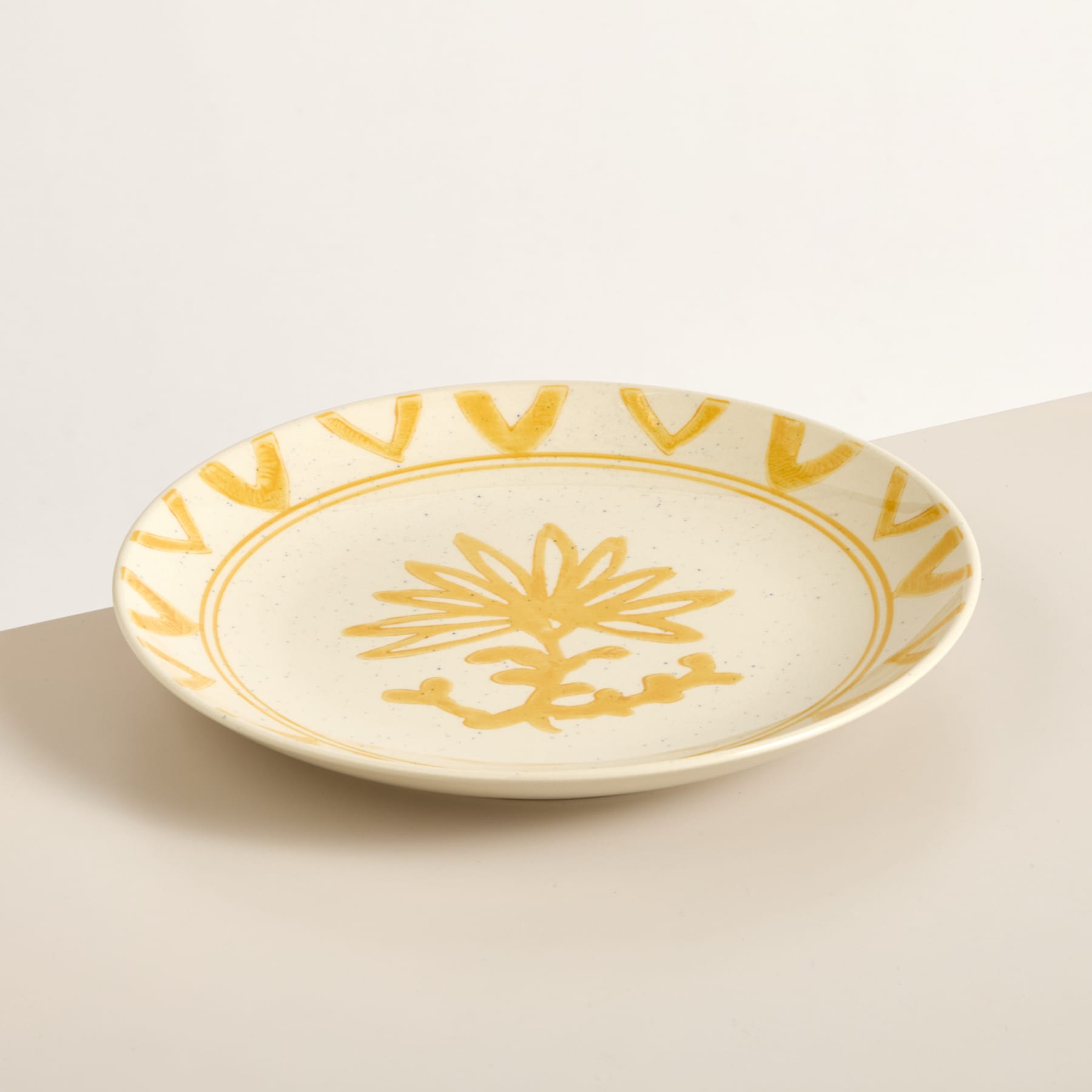 Eine flache Keramikschale mit gelbem Blumenmuster auf cremefarbenem Hintergrund steht auf einer hellen Oberfläche.