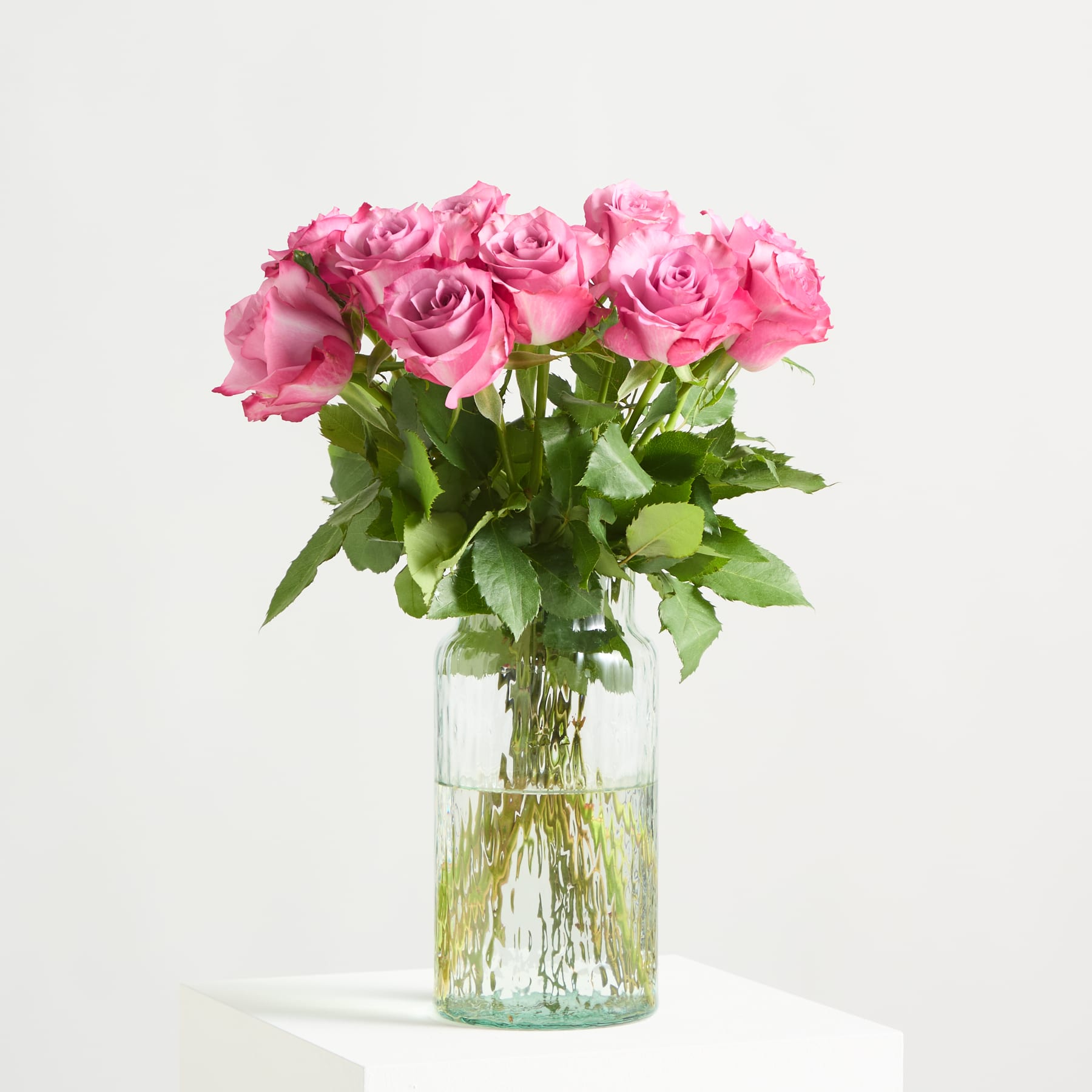 Ein Strauß leuchtend rosa Rosen in einer Glasvase steht vor einem schlichten weißen Hintergrund.