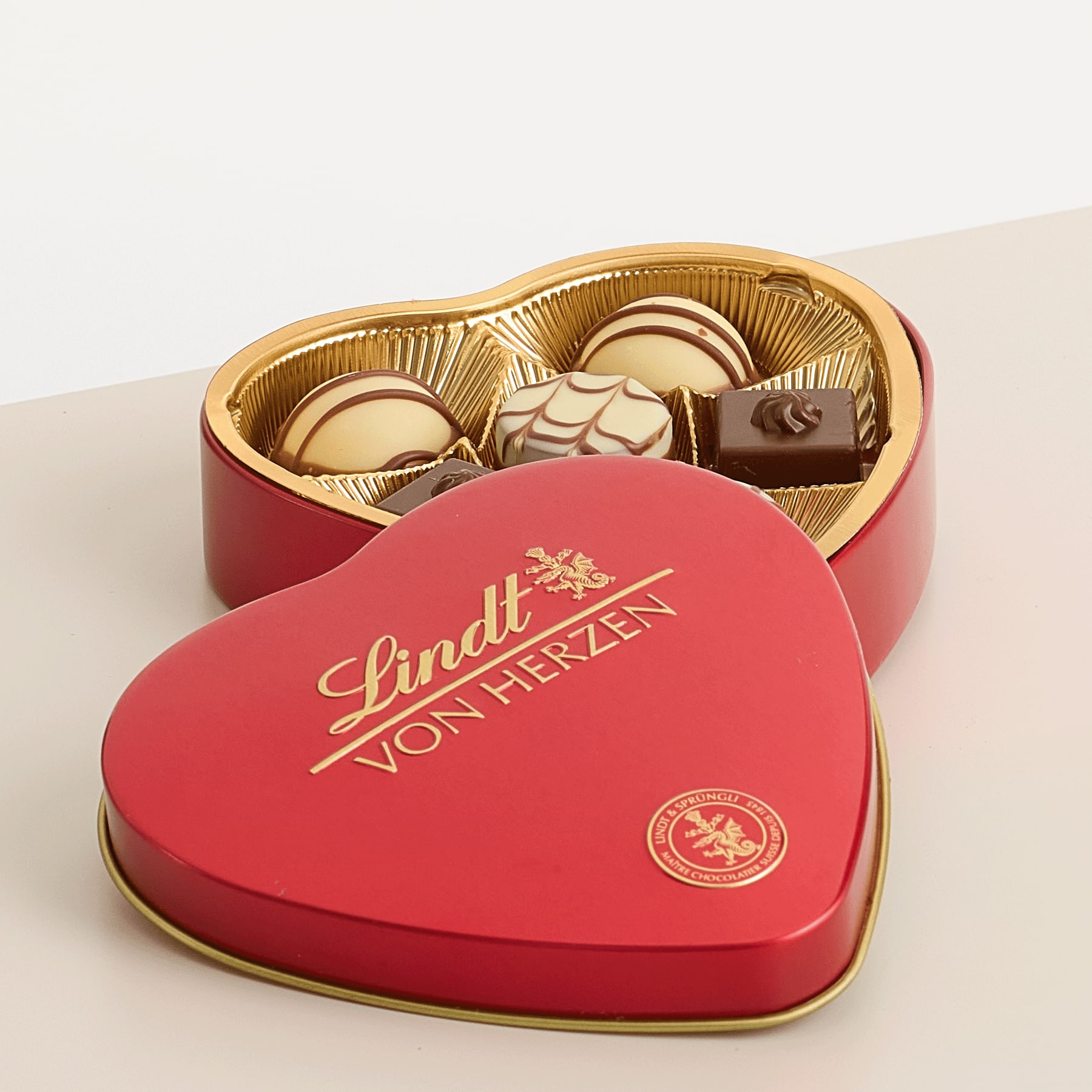 Eine herzförmige rote Schachtel mit einer Auswahl an Lindt-Schokolade vor einem schlichten Hintergrund.