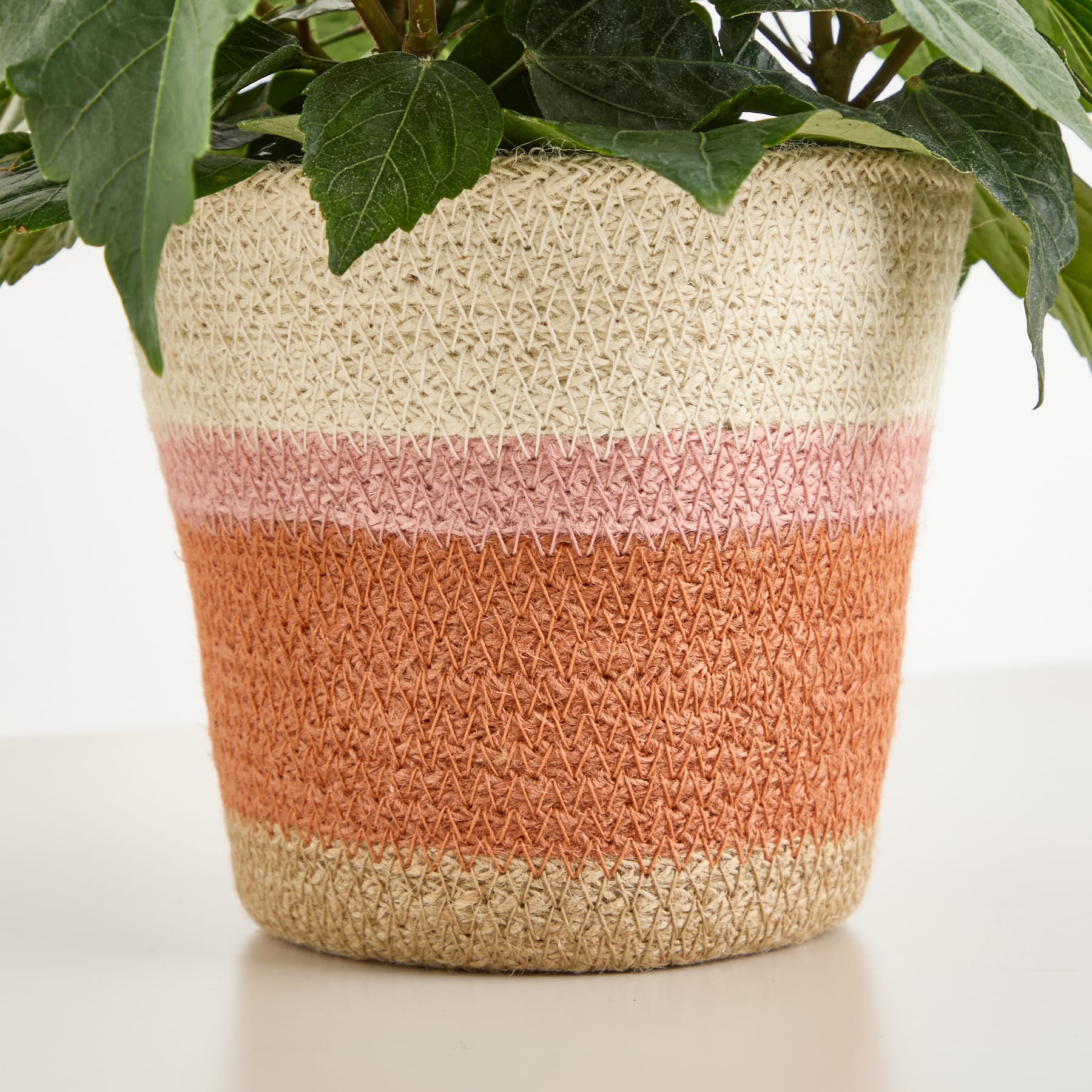 Ein geflochtener Korb oder Pflanztopf mit einem Streifenmuster in Beige-, Rosa- und Orangetönen, im Hintergrund grüne Blätter.