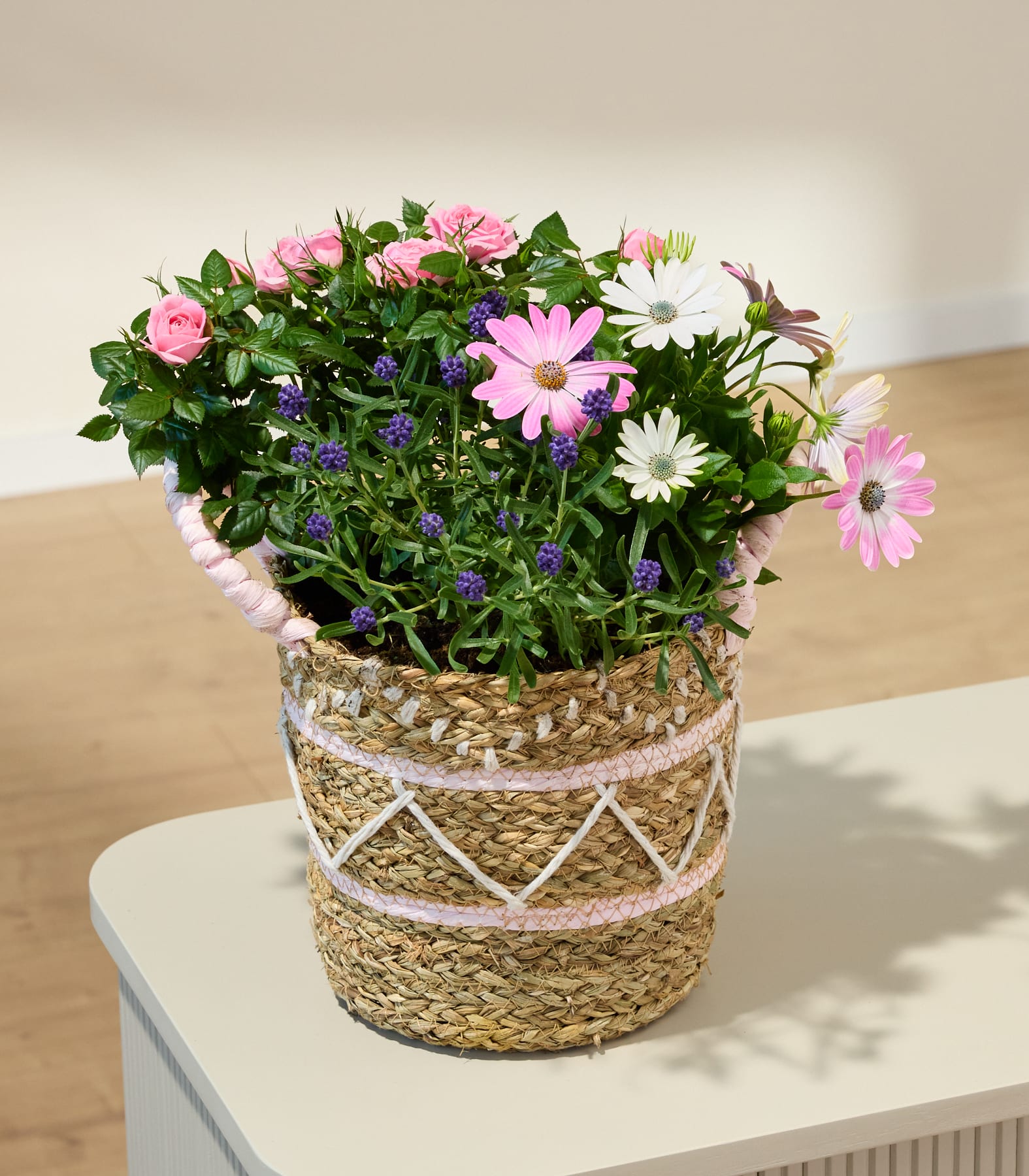 Ein lebendiger Strauß aus rosa Rosen, lila Gänseblümchen und anderen bunten Blumen, arrangiert in einem geflochtenen Korb vor einem schlichten weißen Hintergrund.