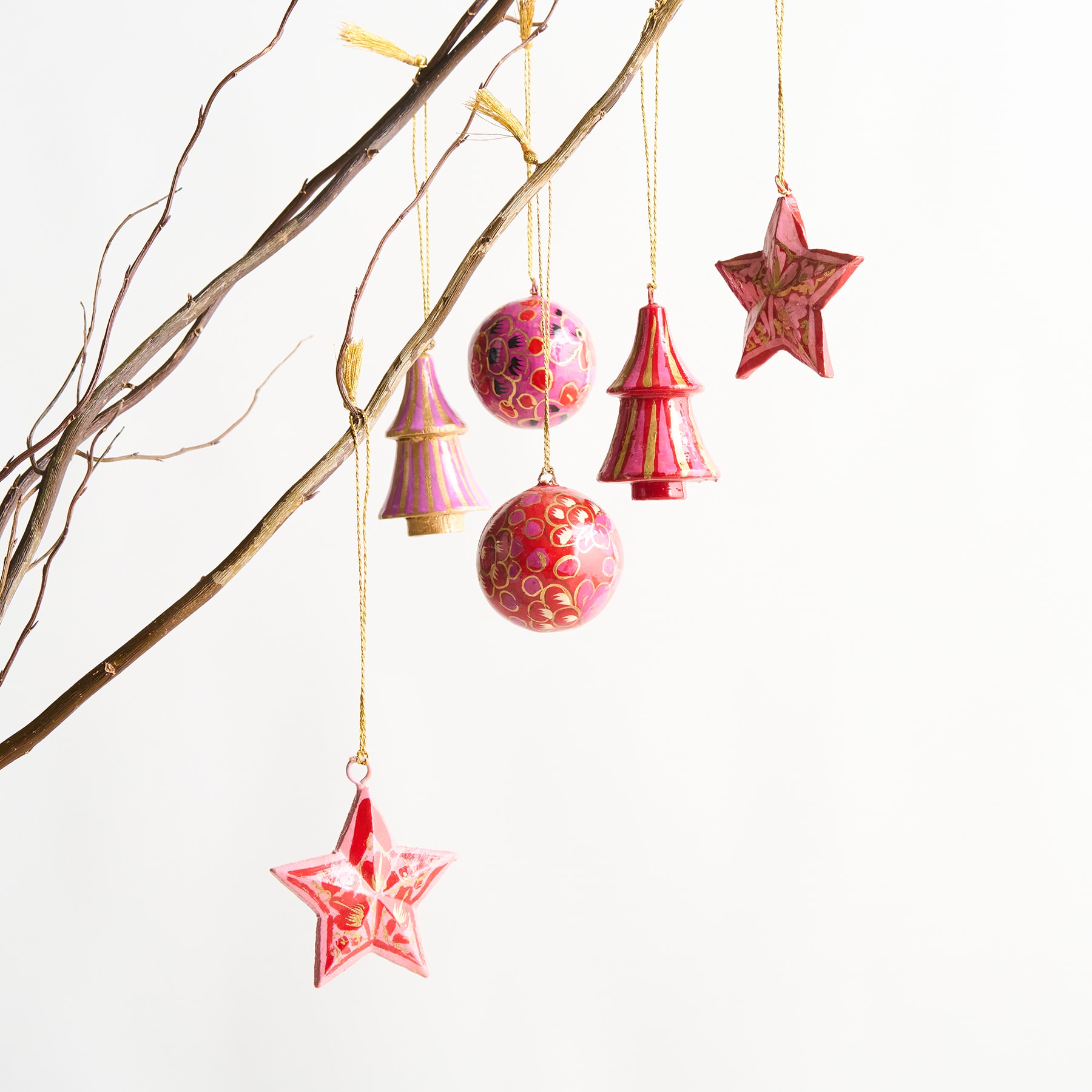 Ein Zweig mit verschiedenen hängenden Ornamenten, darunter rosa und rote Kugeln, Sterne und baumförmige Dekorationen vor einem schlichten weißen Hintergrund.