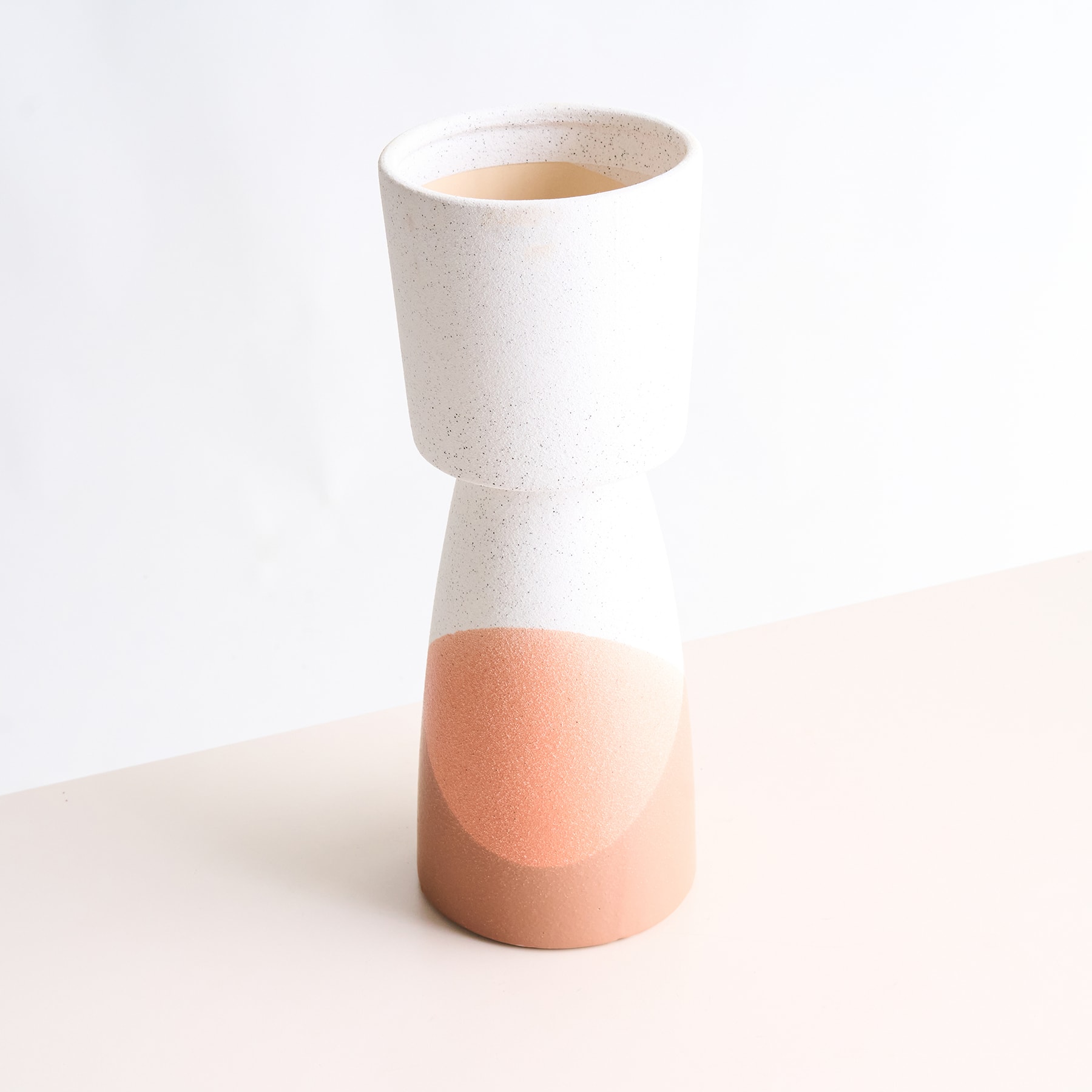 Eine weiße Keramikvase mit pfirsichfarbenem Sockel steht auf einer hellen Oberfläche vor einem schlichten weißen Hintergrund.