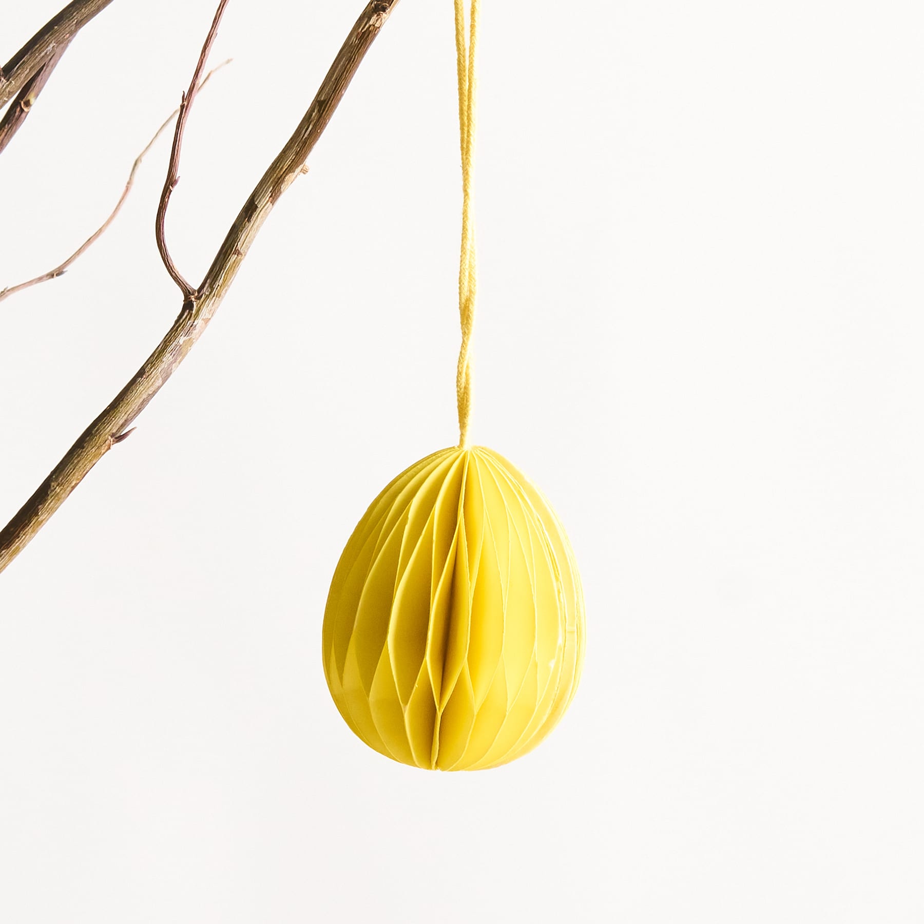 Ein leuchtend gelbes Papierornament mit Wabenmuster hängt vor einem schlichten weißen Hintergrund an einem Ast.