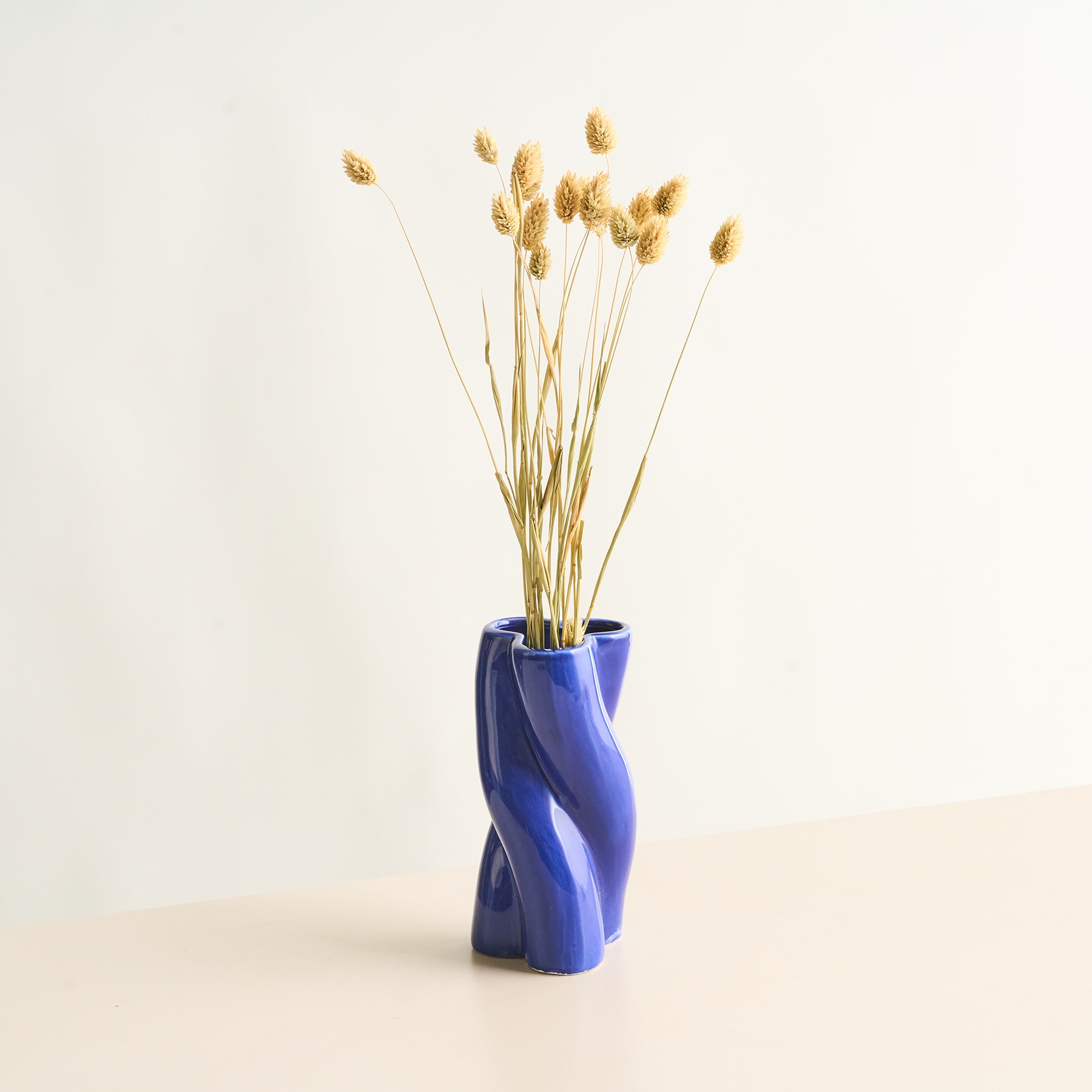Eine leuchtend blaue Keramikvase enthält einen Strauß zarter, goldfarbener Trockenblumen vor einem schlichten, hellen Hintergrund.