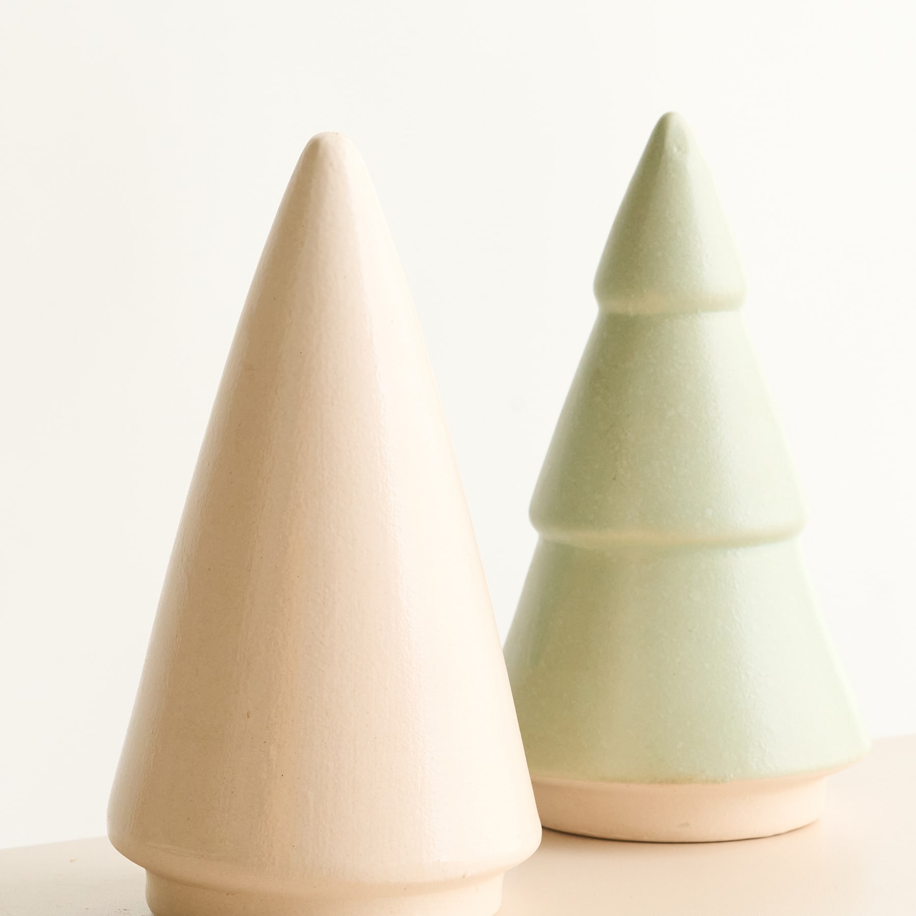 Vor einem schlichten weißen Hintergrund stehen zwei minimalistische Weihnachtsbäume aus Keramik in verschiedenen Beige- und Grüntönen.