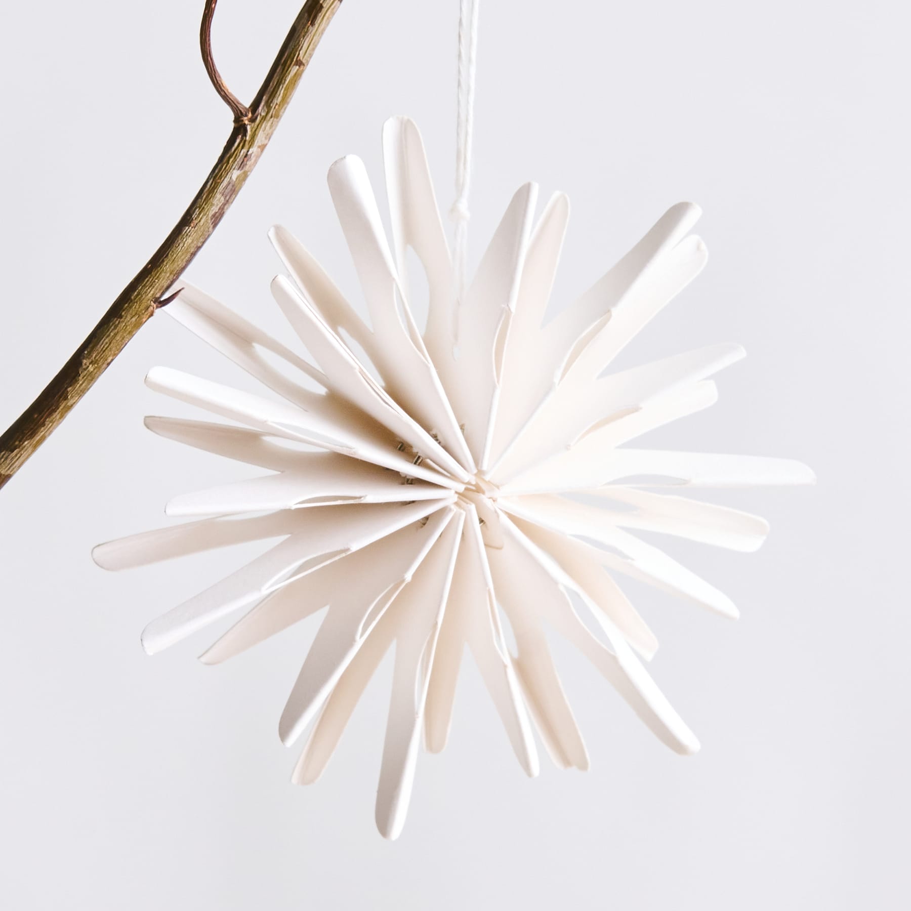 Ein zartes, sternförmiges Ornament aus weißem Papier hängt vor einem schlichten weißen Hintergrund an einem Holzast.