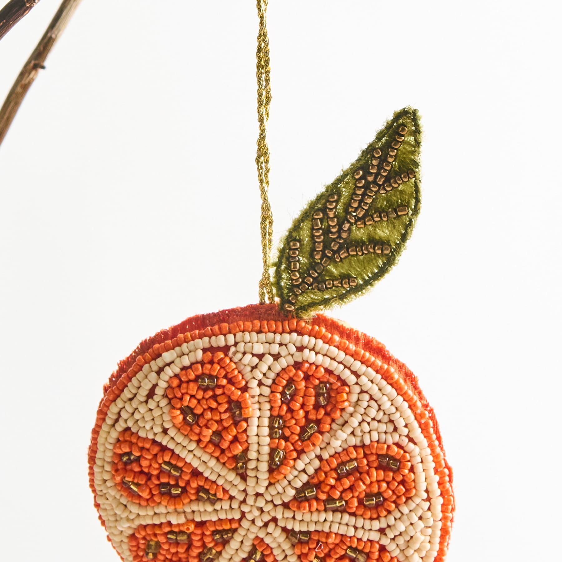 Ein farbenfrohes Perlenornament in Form einer Zitrusfrucht mit komplizierten Mustern und einem goldenen, blattähnlichen Element hängt vor einem schlichten weißen Hintergrund.