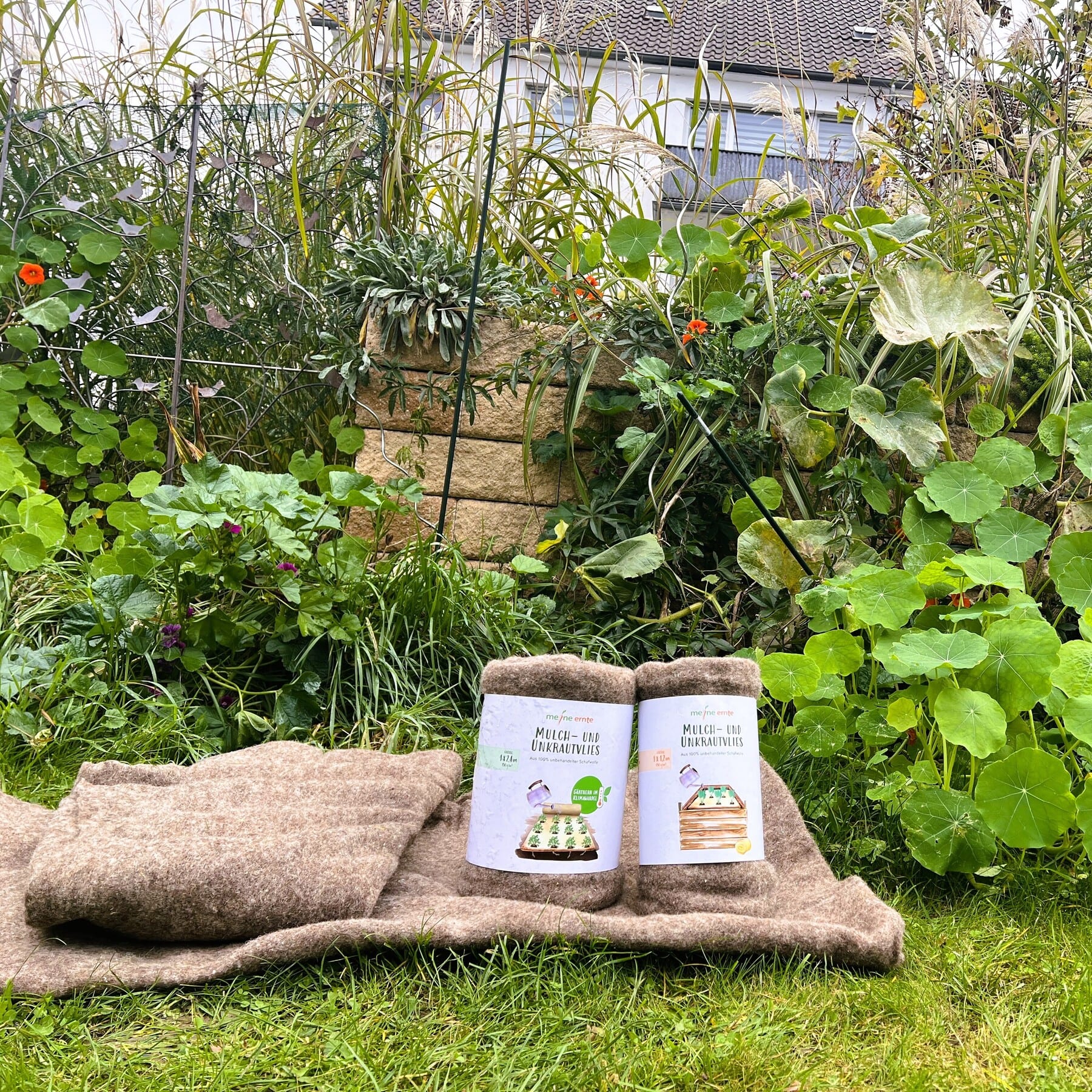 Im Vordergrund sind zwei Taschen mit Gartenbedarf auf einem Sackleinen zu sehen, umgeben von einem üppigen, verwilderten Garten mit verschiedenen Pflanzen und Vegetation im Hintergrund.