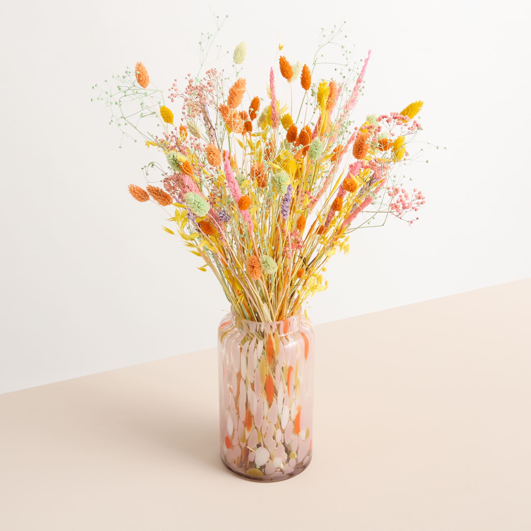Ein farbenfroher Strauß getrockneter Blumen in verschiedenen warmen Farbtönen, darunter Orange, Gelb und Rosa, wird in einer Glasvase vor einem schlichten, hellen Hintergrund präsentiert.