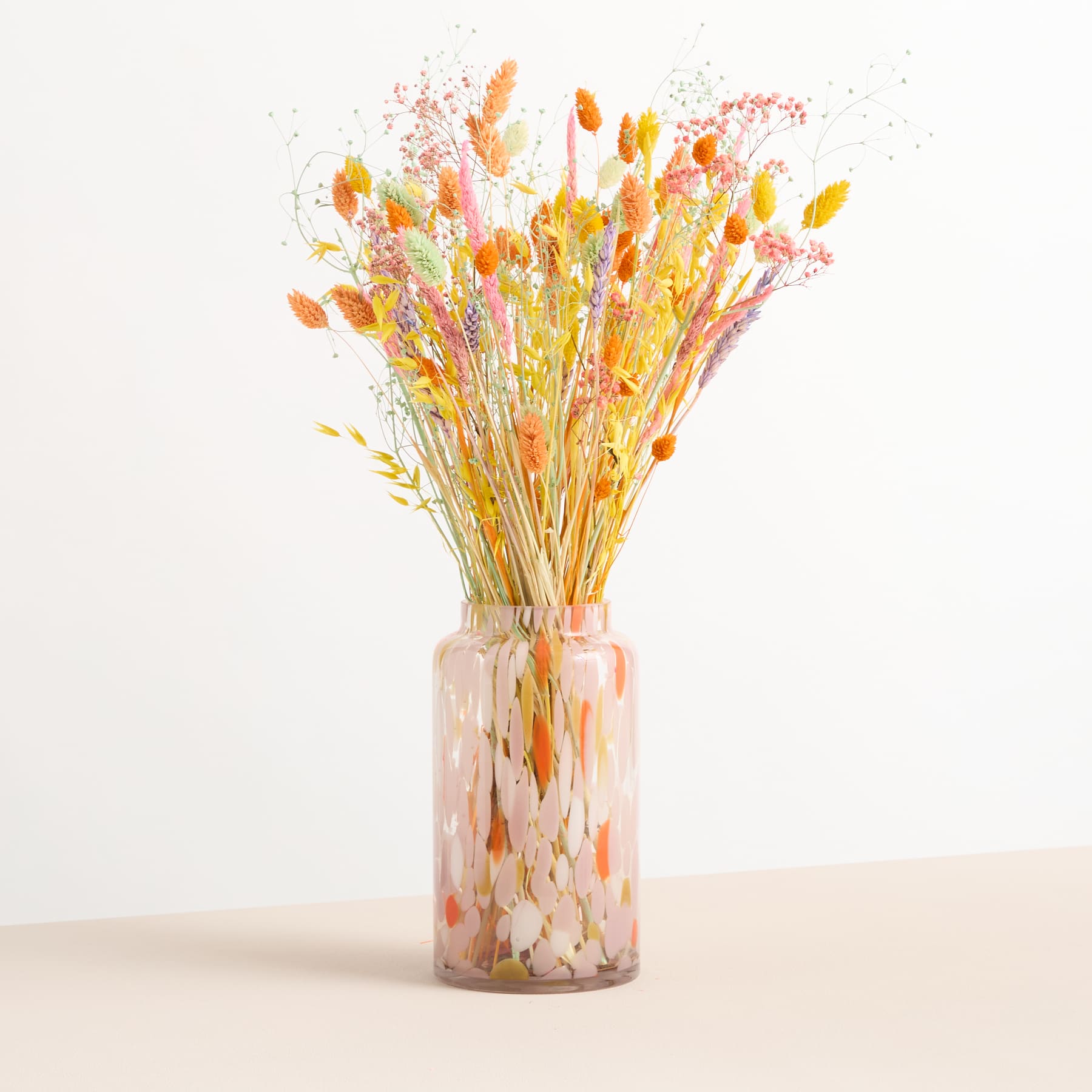 Ein farbenfroher Strauß getrockneter Blumen in warmen Herbstfarben, darunter Gelb-, Orange- und Rottöne, wird in einer Glasvase vor einem schlichten weißen Hintergrund präsentiert.