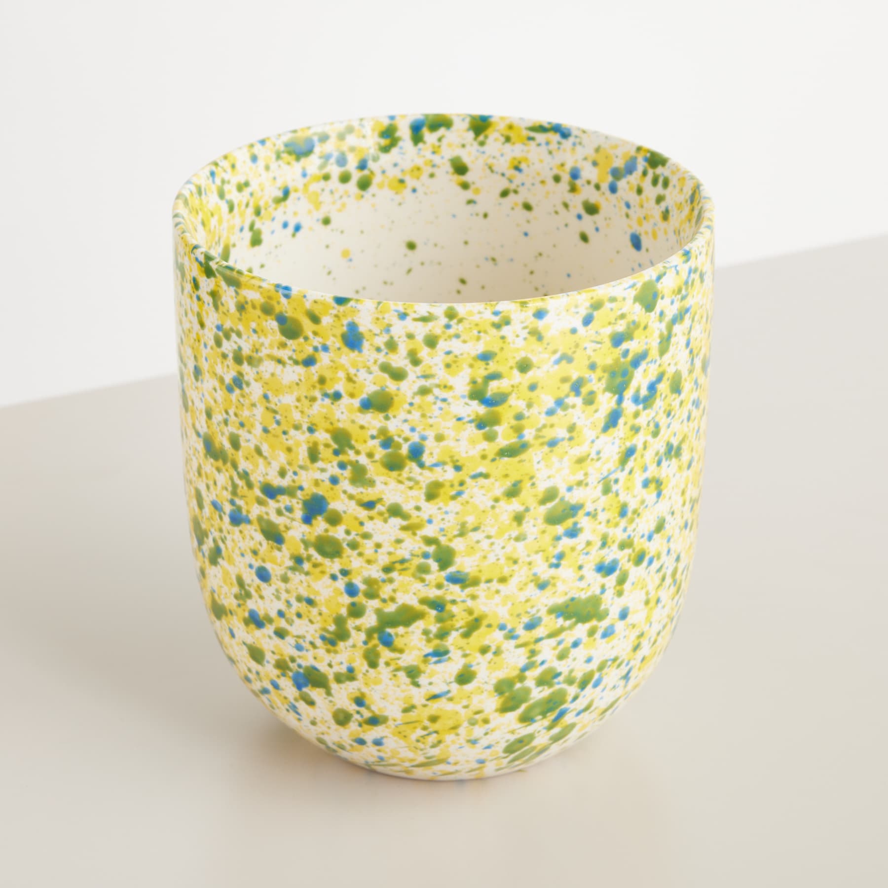 Eine Tasse oder ein Gefäß aus Keramik mit einem gesprenkelten Muster aus gelben, grünen und blauen Punkten oder Spritzern auf einem hellen Hintergrund.