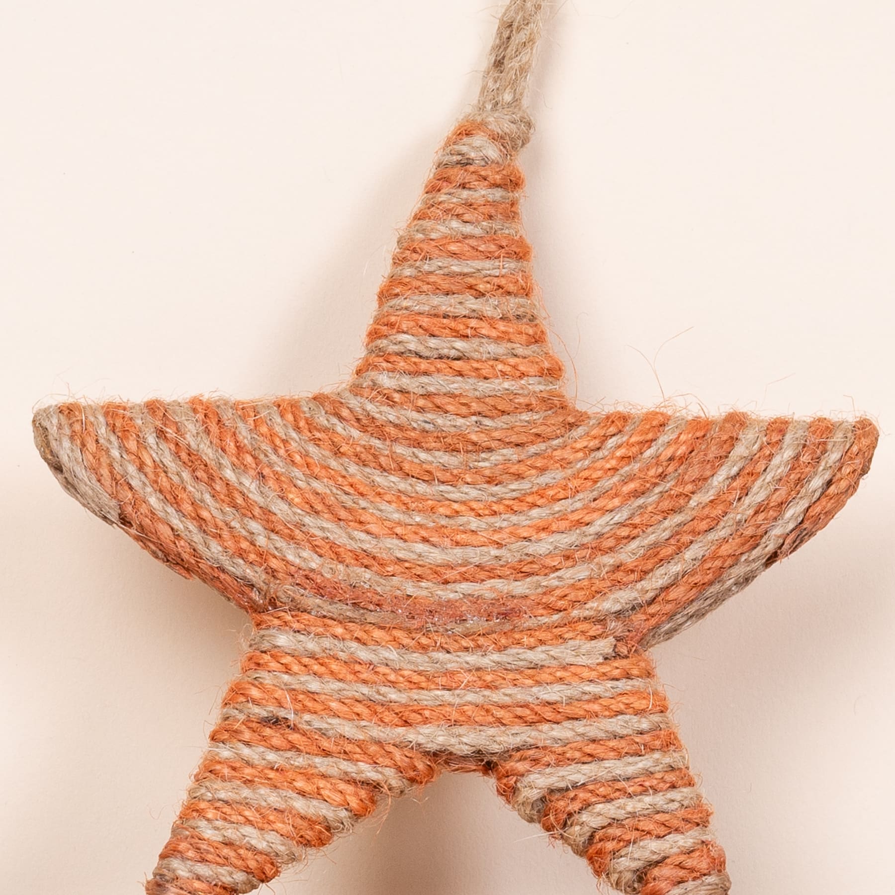 Ein gewebtes sternförmiges Ornament aus orangefarbenen und beigen Seilen auf einem schlichten, hellen Hintergrund.
