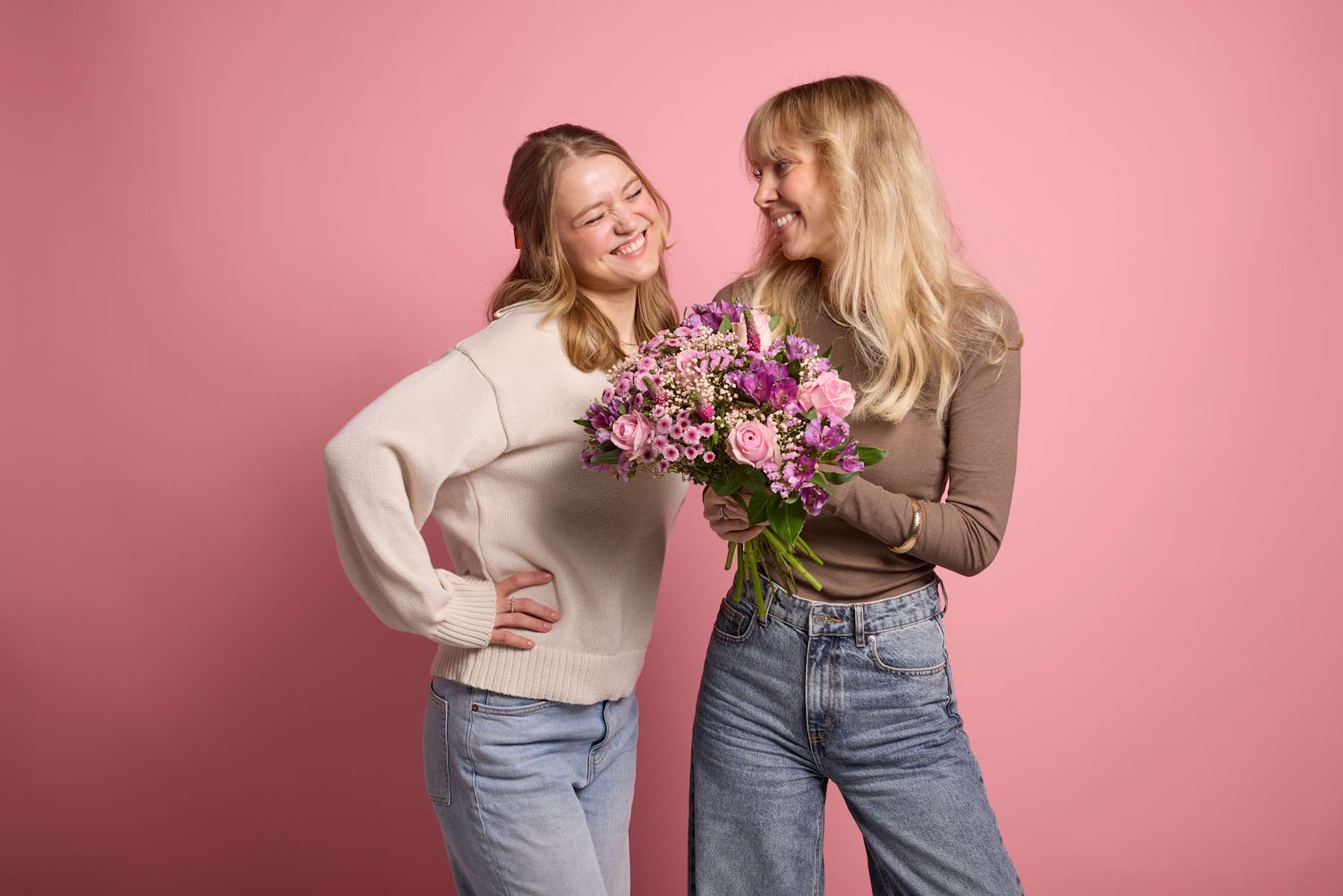 Zwei junge Frauen stehen zusammen, eine hält einen Strauß lila Blumen vor einem rosa Hintergrund.