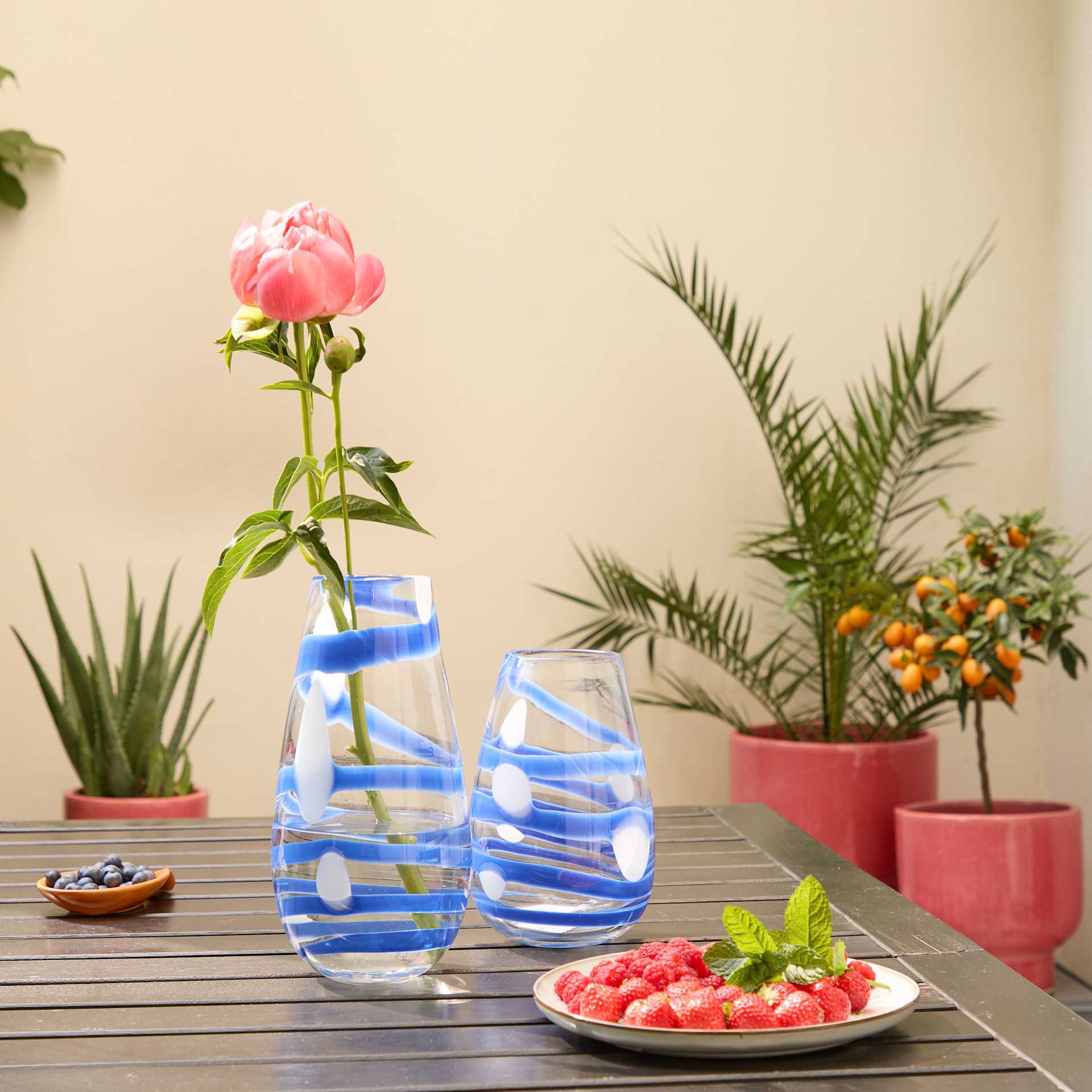 Eine rosa Pfingstrosenblüte in einer blau-weiß gemusterten Vase steht auf einem Holztisch, umgeben von Topfpflanzen und einem Teller mit frischen Erdbeeren.