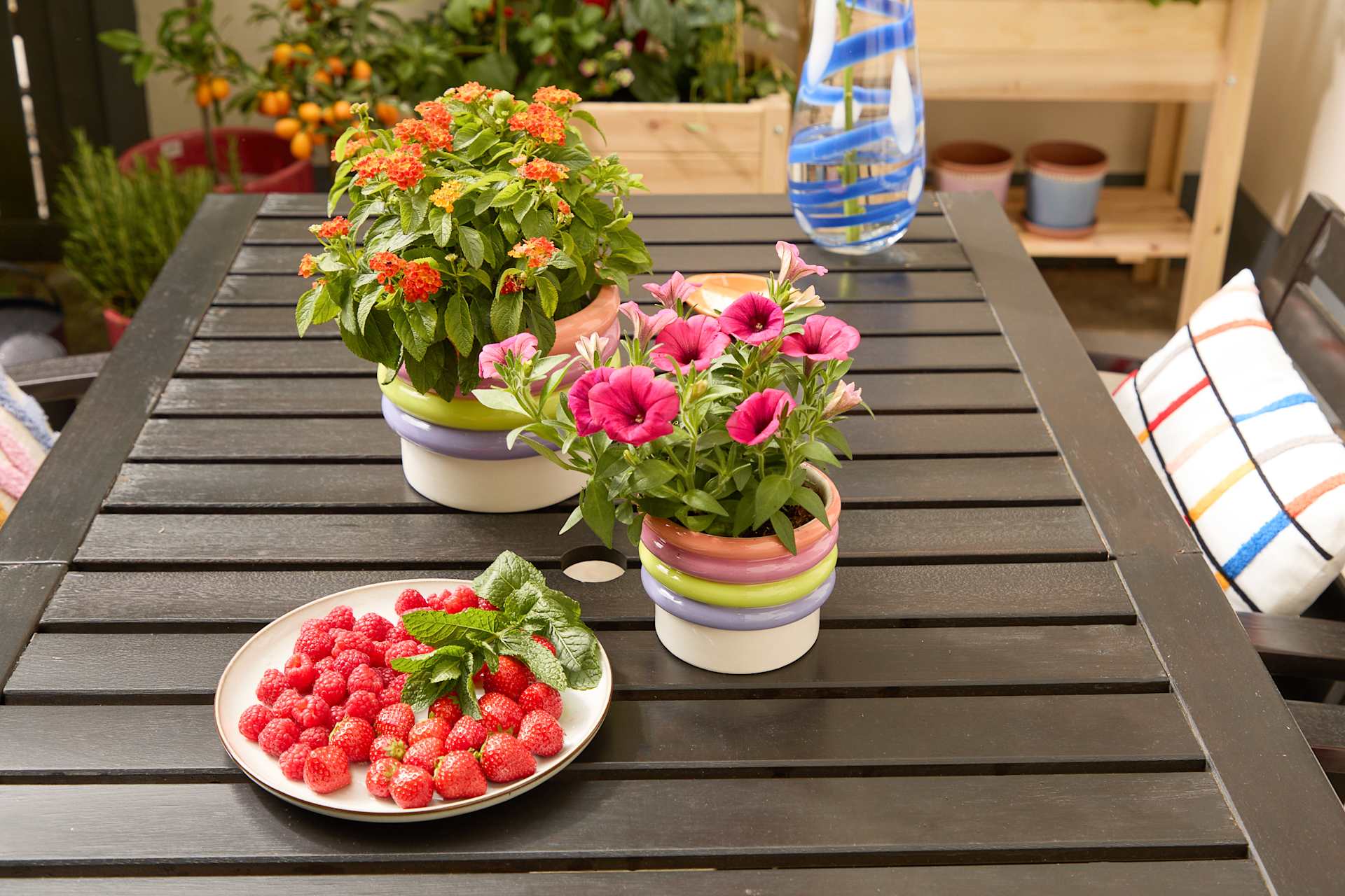 Ein Holztisch im Freien, geschmückt mit leuchtend rosa Blumen in gemusterten Vasen und einem Teller mit frischen, reifen Erdbeeren.