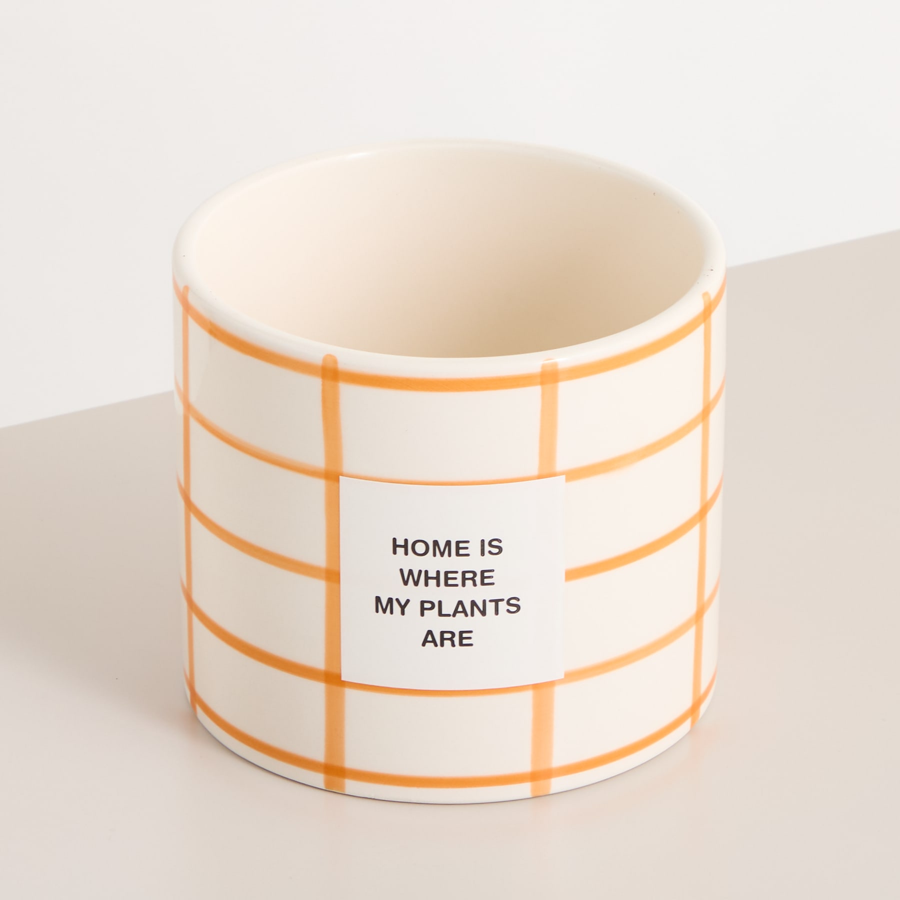Eine Keramiktasse mit einem orangefarbenen Gittermuster und dem Aufdruck „HOME IS WHERE MY PLANTS ARE“ steht auf einer hellen Oberfläche.