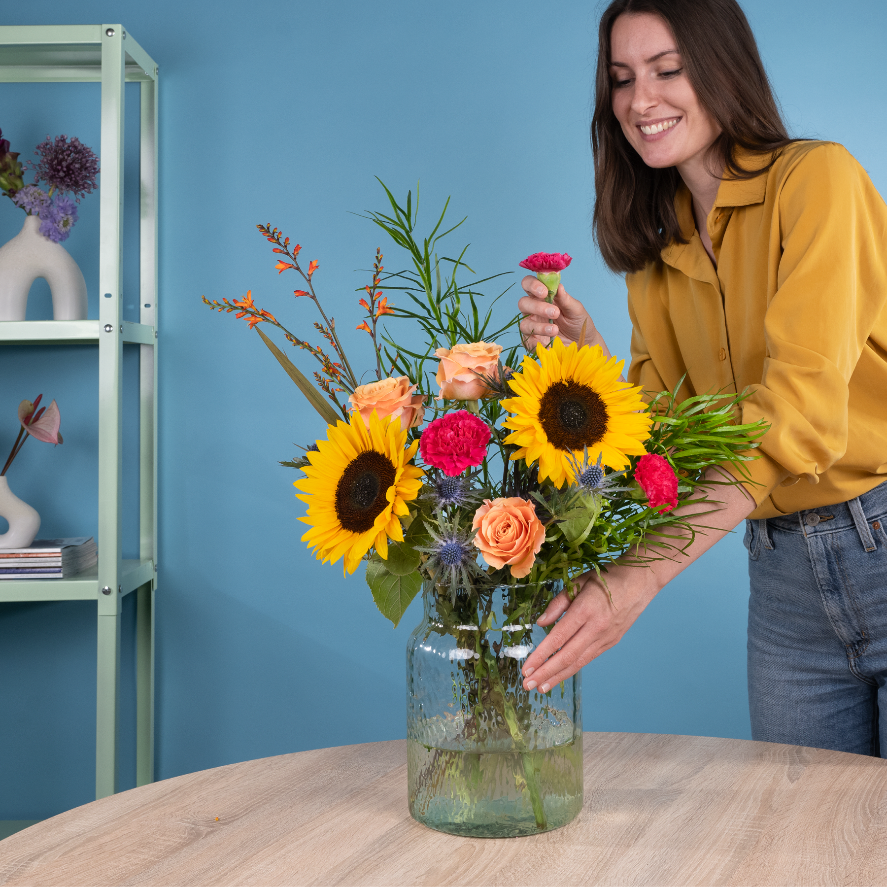 Eine lächelnde Frau in einer gelben Jacke hält einen farbenfrohen Strauß aus Sonnenblumen, Rosen und anderen bunten Blumen vor einer blauen Wand mit Regalen im Hintergrund.